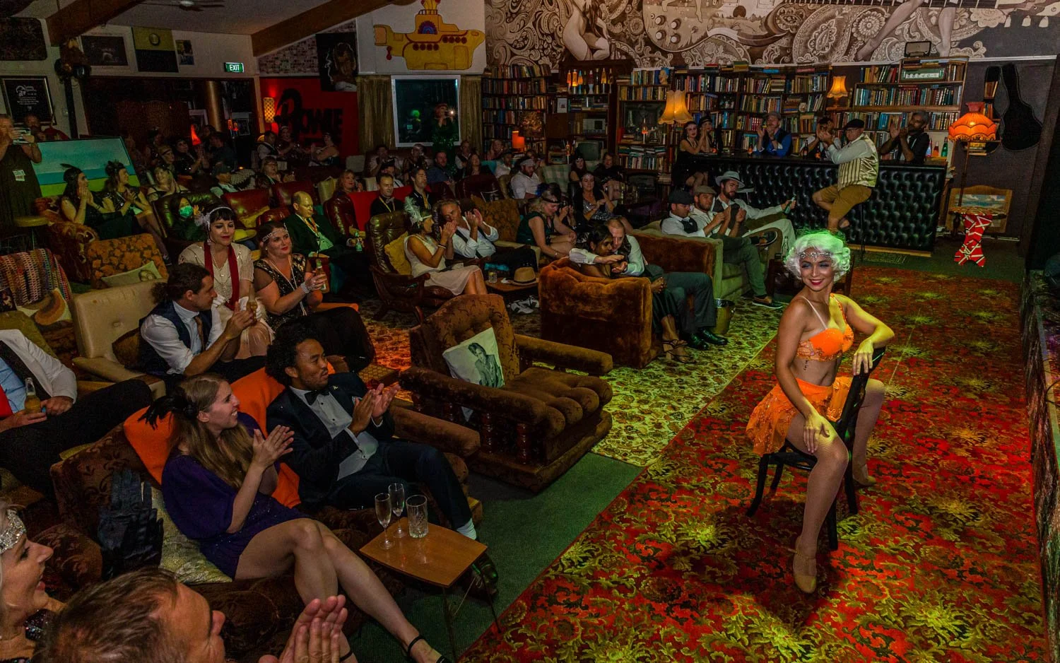 A burlesque performer in an orange costume posing on a chair in front of an audience at a club with eclectic decor and a patterned carpet.