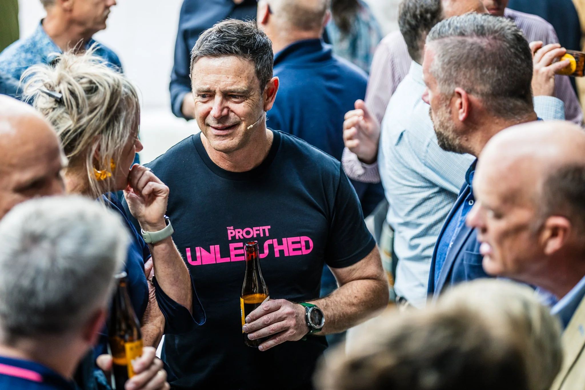 A group of people at a social gathering, with a man in the center wearing a black t-shirt that reads 'The Profit Unleashed' in pink, holding a beer bottle and talking to a woman with blonde hair and earrings.