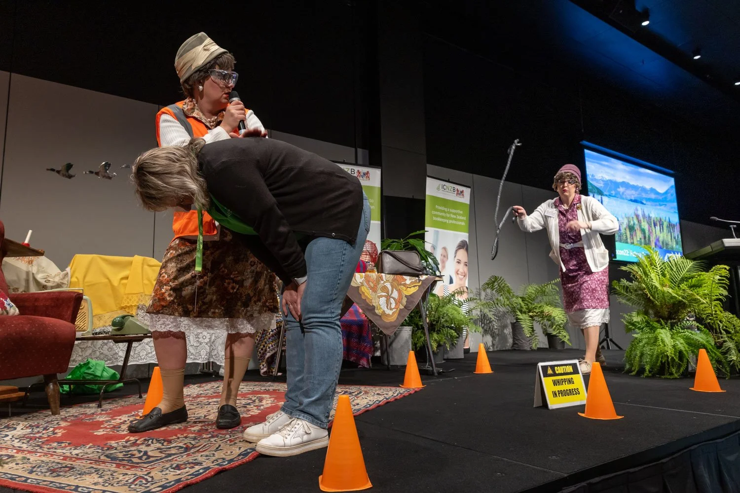 Two women performing a comedy skit on stage, one horizontal with her head down and the other standing holding a whip, with props and potted plants around, and a large screen in the background.