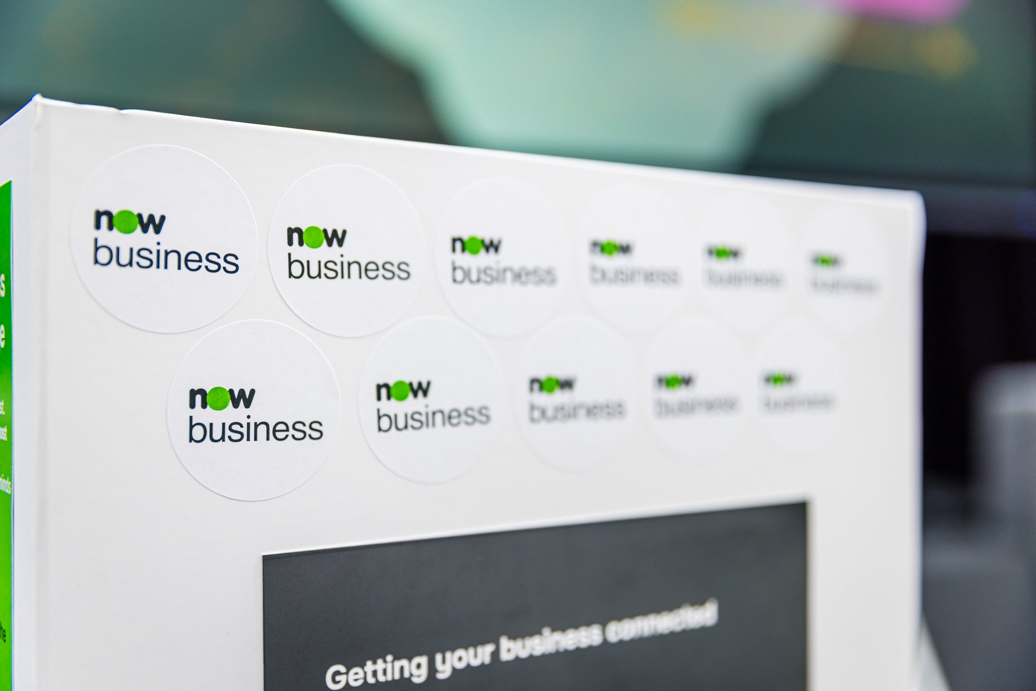 Close-up of a white retail display box with multiple stickers of the 'now business' logo, featuring the word 'now' in black and green and 'business' in black, arranged in a grid pattern.