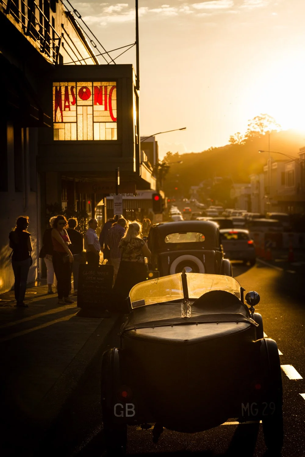 People standing in line on a city sidewalk during sunset, vintage cars parked along the street, and a building with a stained-glass window that says 'HISTORIC'.