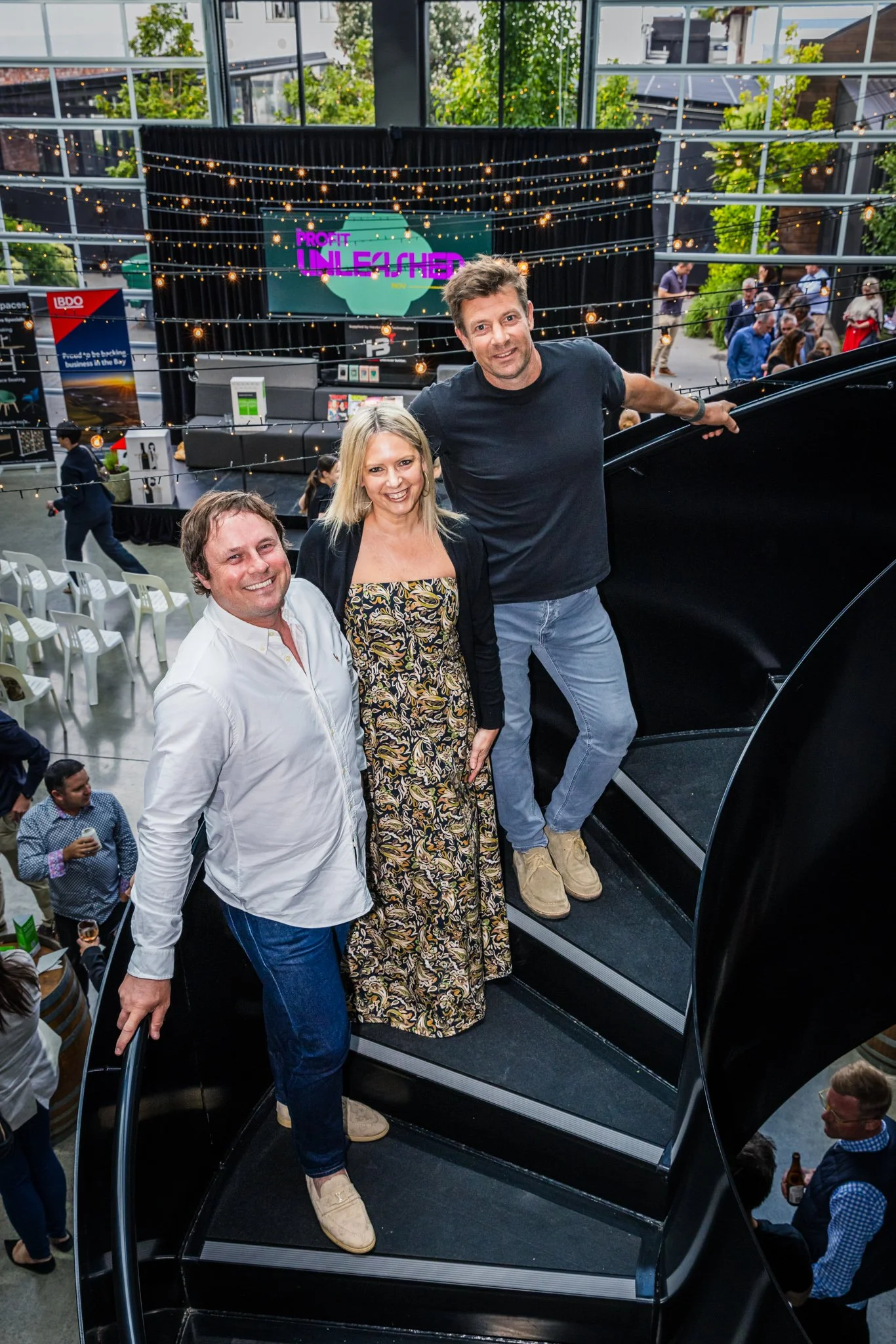 Three people smiling on a staircase at an indoor event with a stage in the background, balloons, and a crowd of people, Some of whom are holding drinks.