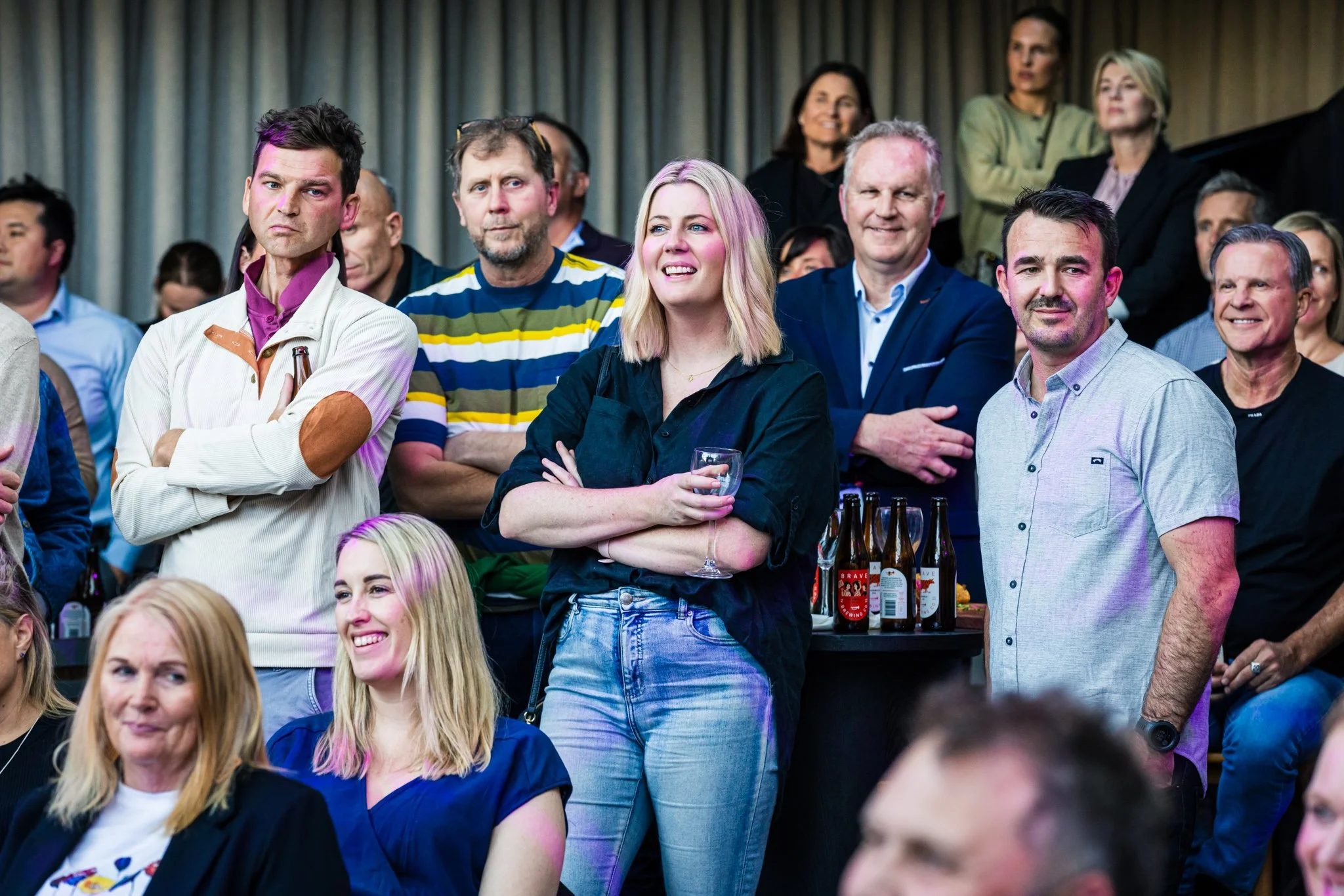 Group of people attending a social event, standing and sitting, with some holding drinks, indoors with curtains in the background.