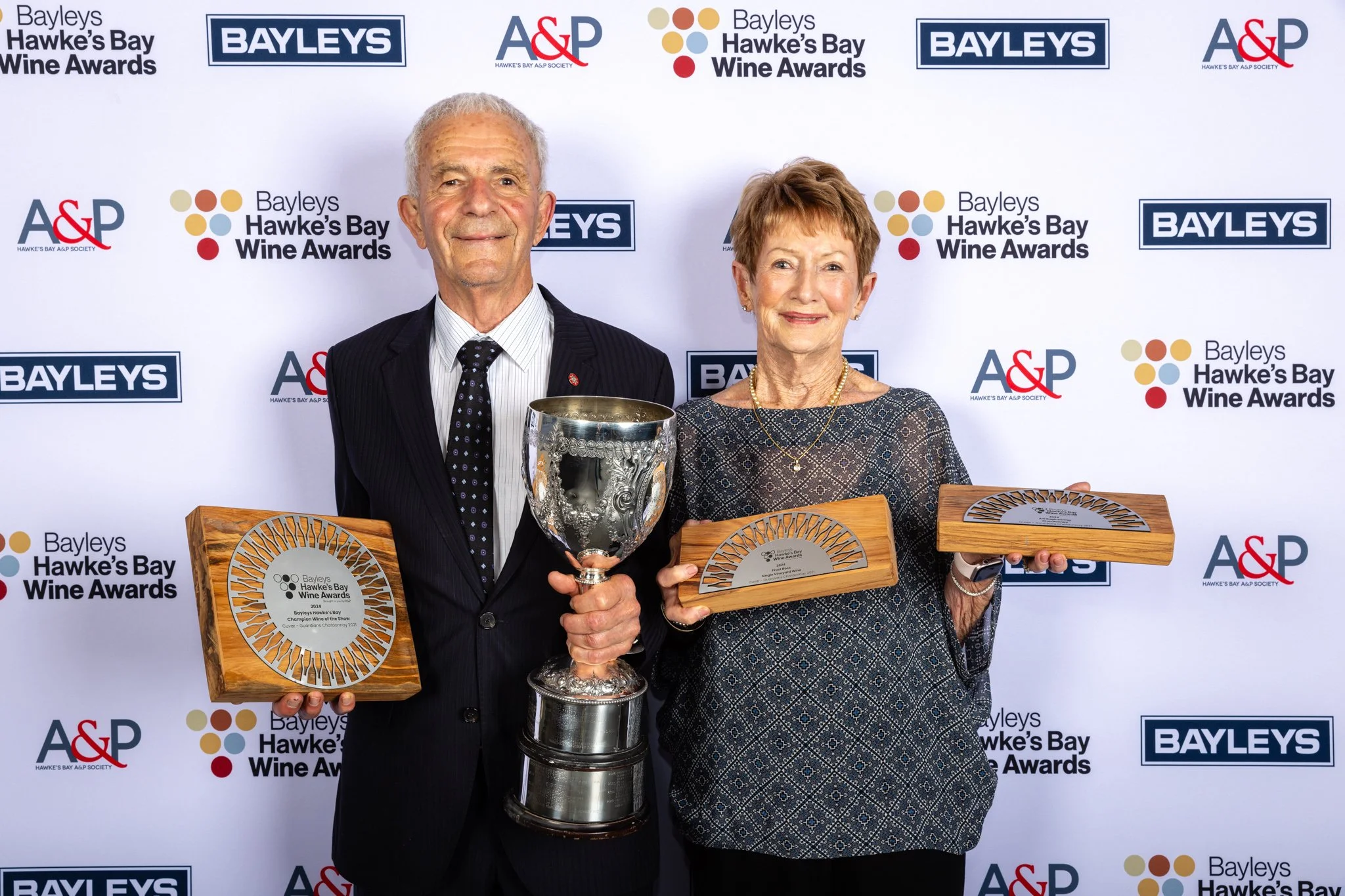 An elderly man and woman standing together at an awards ceremony, holding plaques and a trophy, in front of a backdrop with logos for Bayleys, A&P, and Hawke's Bay Wine Awards.