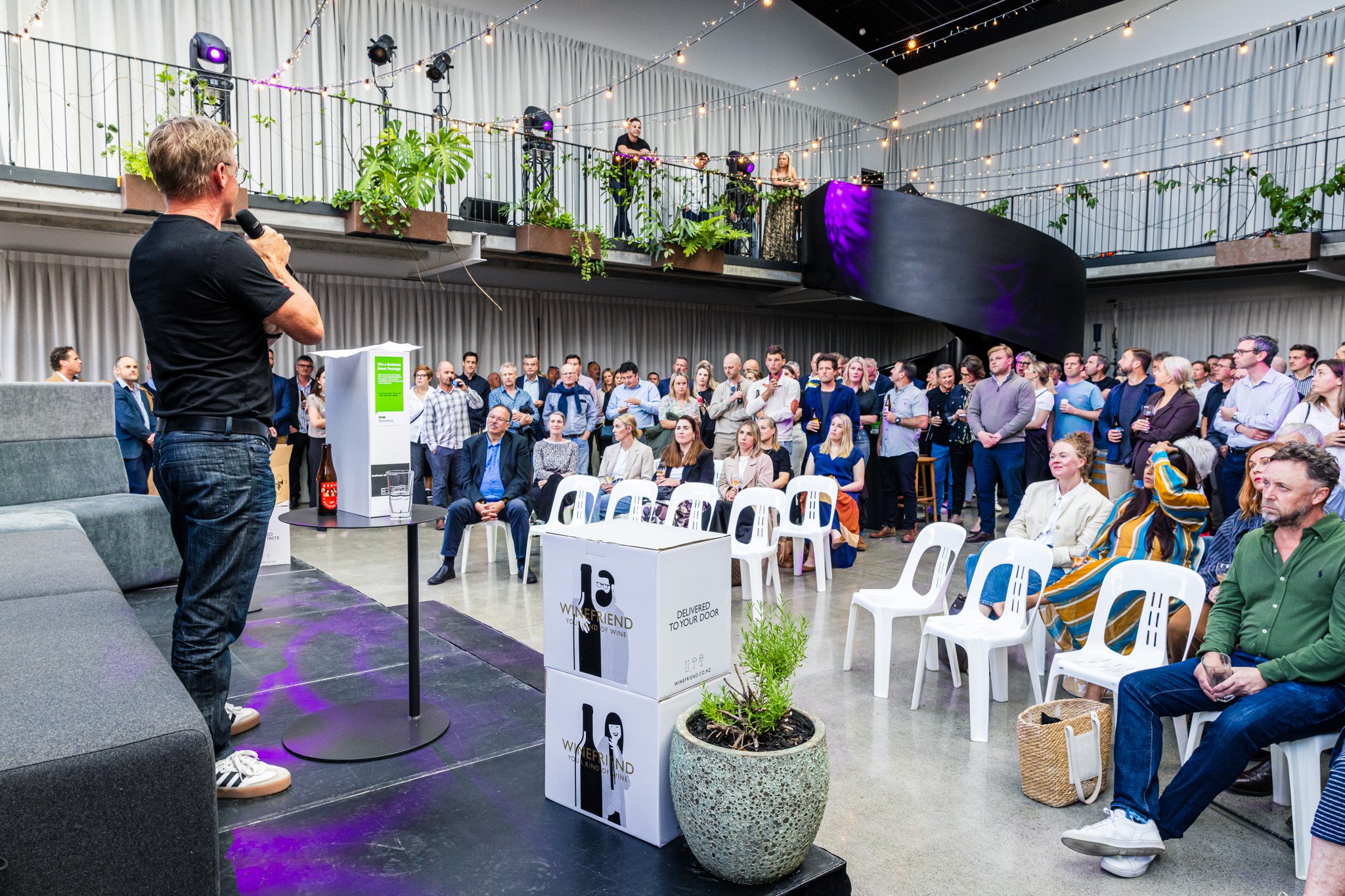 A speaker in black t-shirt and jeans holding a microphone addresses a seated and standing audience in a large indoor event space decorated with string lights and plants, with a group of DJs or musicians on a balcony in the background.