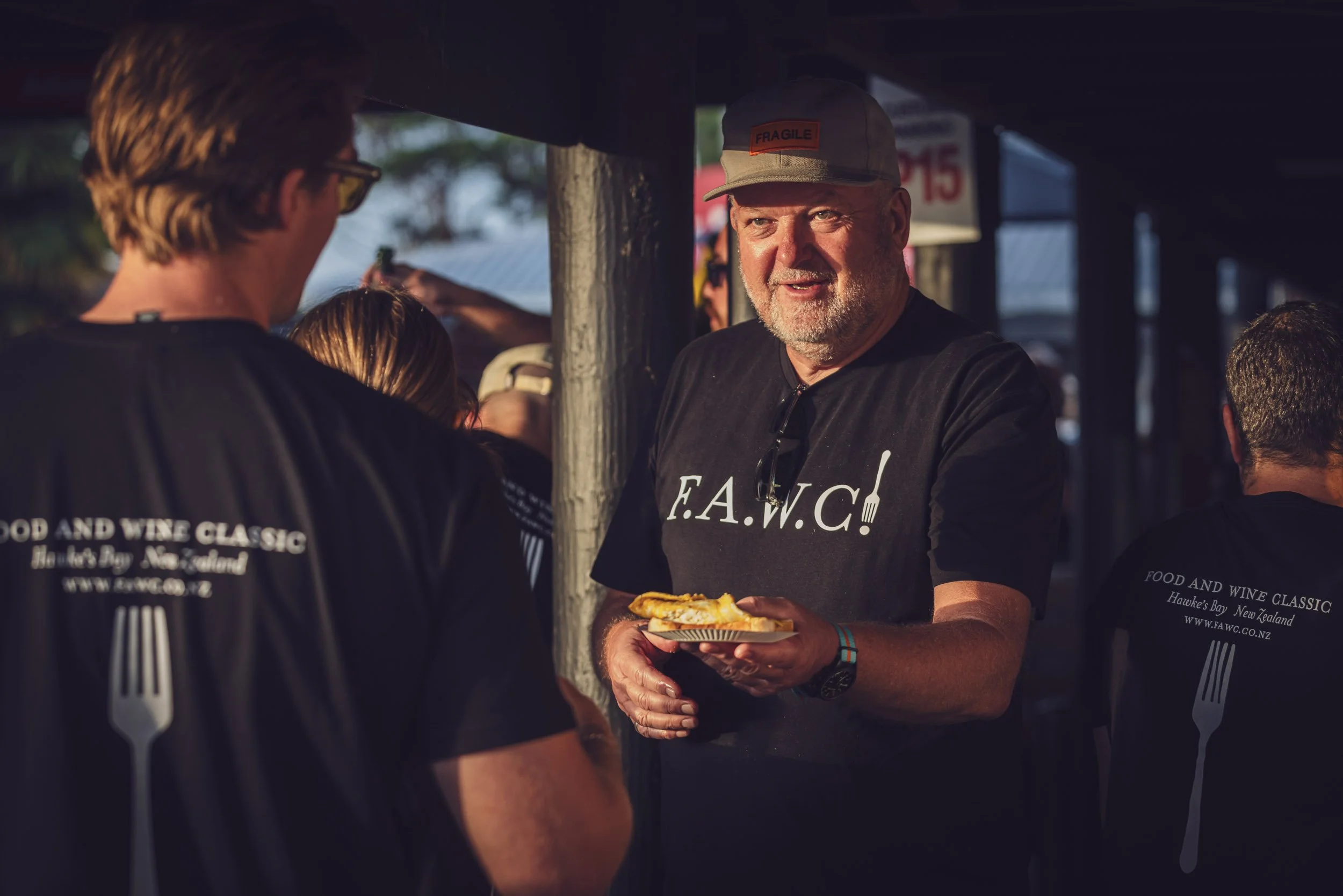 A man with a beard wearing a gray cap and black shirt holding a plate of food, standing and talking to two other people at an outdoor event in the evening.