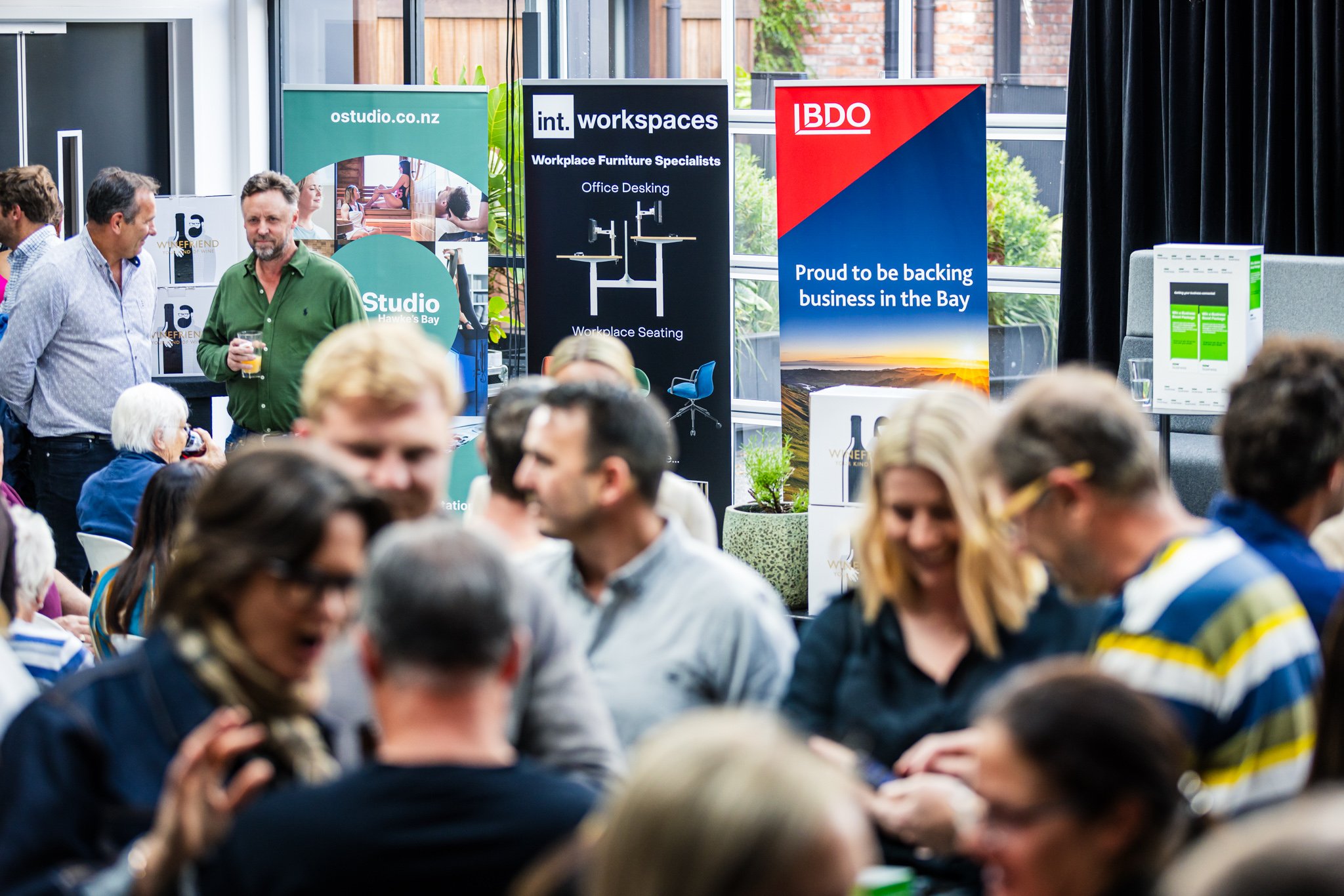A busy networking event or conference with multiple people engaging in conversation, with banners and screens in the background advertising workspace furniture and local businesses.