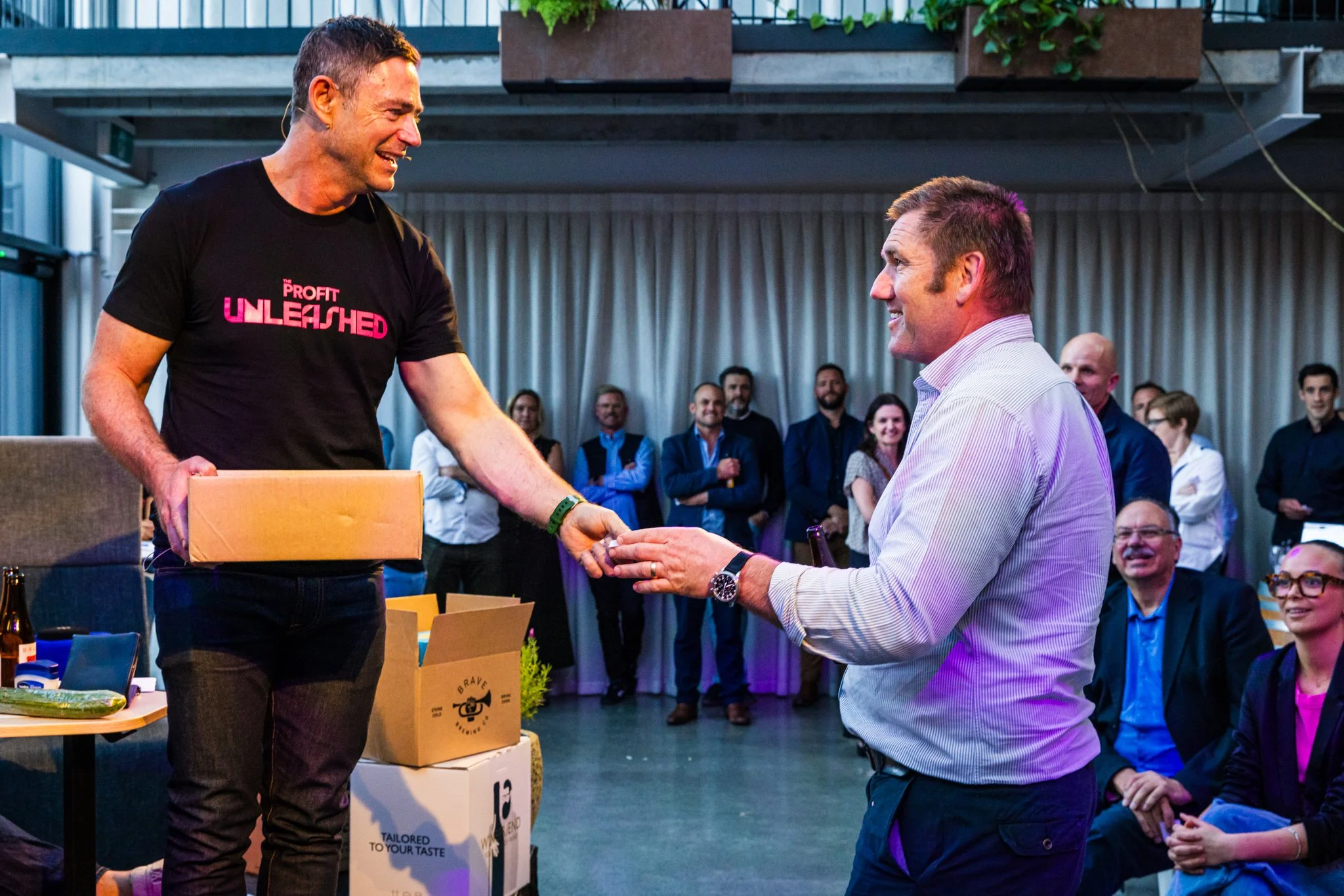 A man in a black t-shirt with pink text, handing over a box to another man in a striped shirt during an indoor event with an audience watching and smiling.