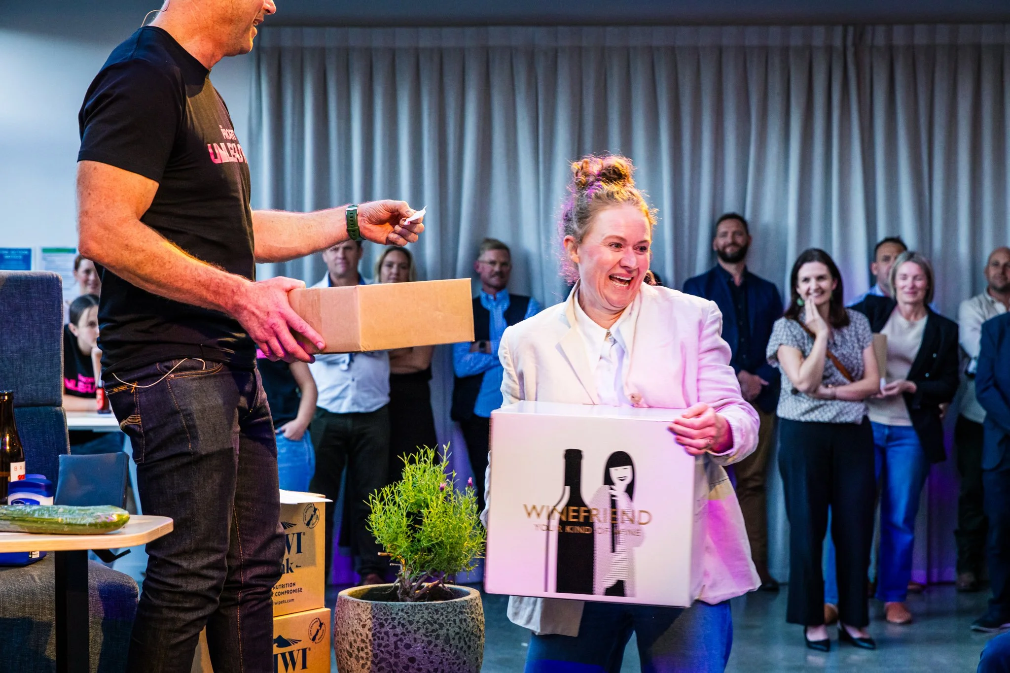 Woman receiving a gift box from a man at a gathering or celebration, with several onlookers smiling and watching in the background.