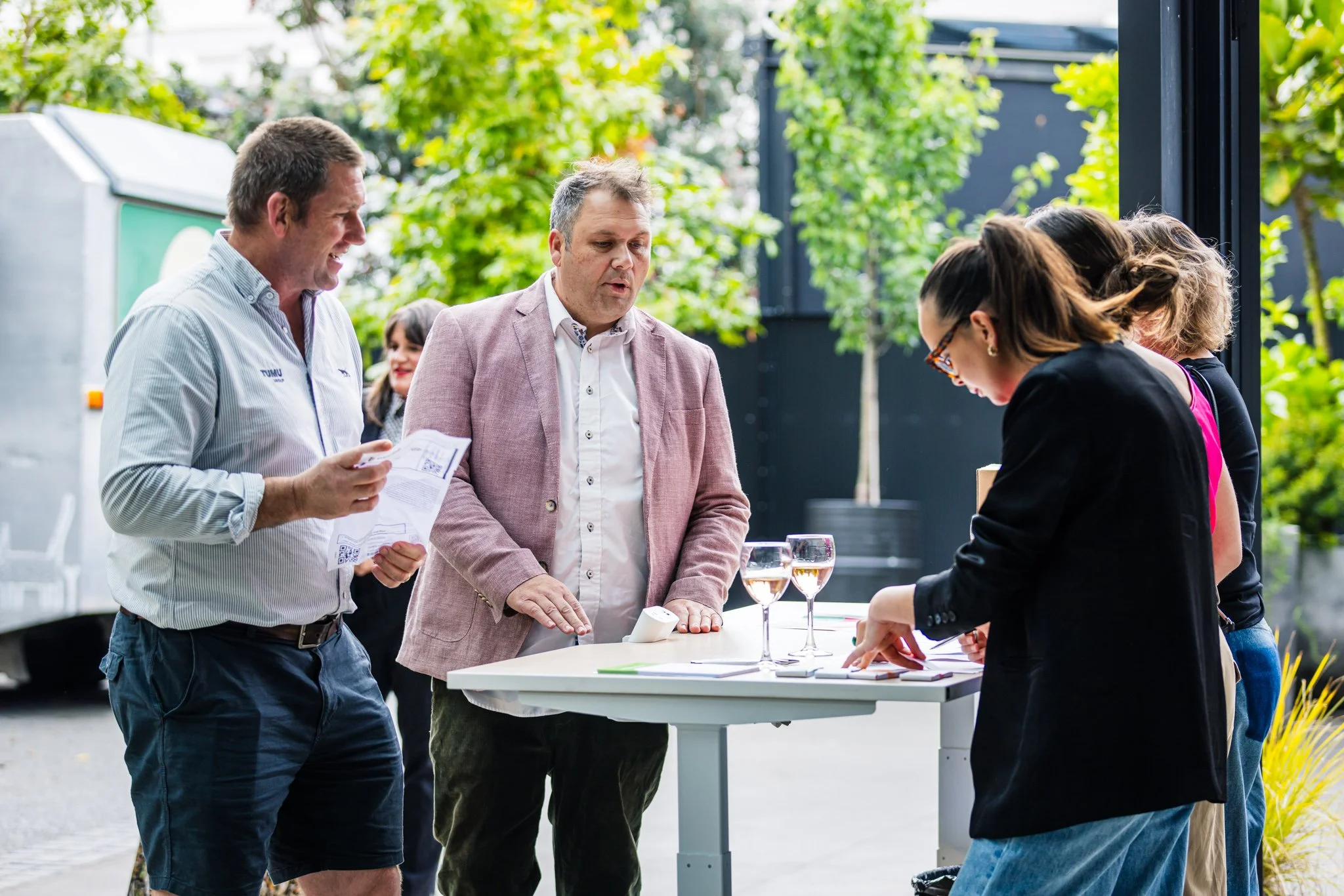 People gathered around a table outdoors, talking and looking at papers, with glasses of rose wine on the table and trees in the background.