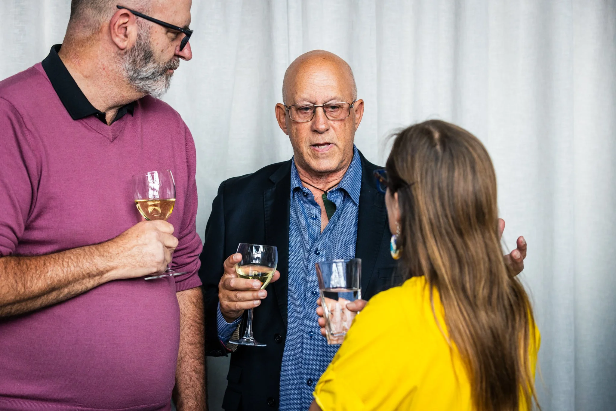 Three people having a conversation at a social event, two men and one woman, with drinks in hand, standing in front of a white curtain.