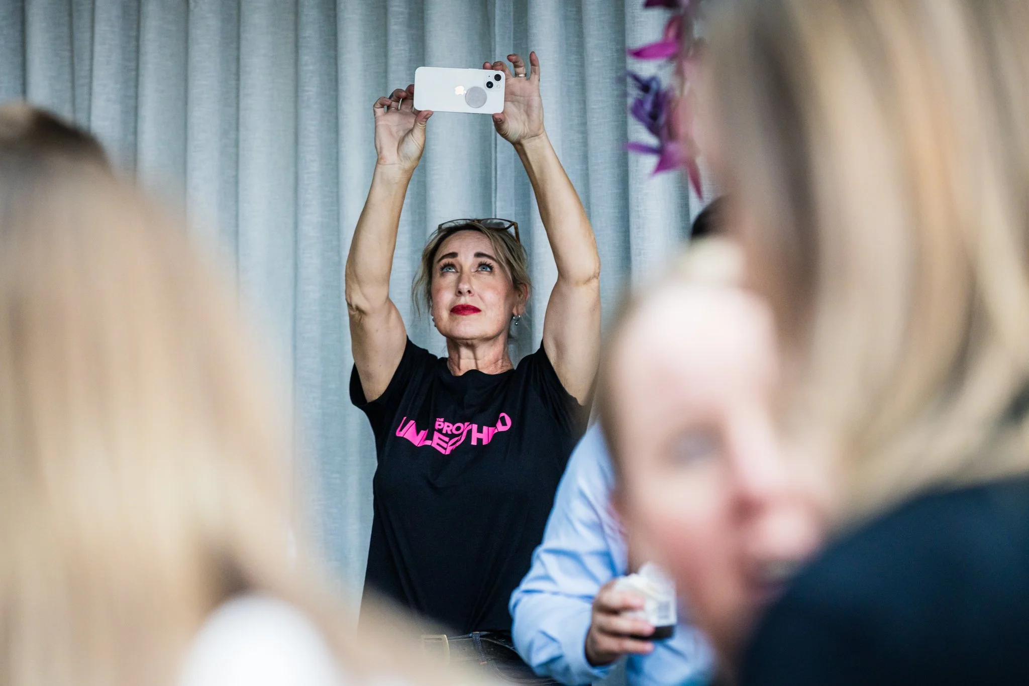 Woman taking a selfie with her smartphone at an indoor gathering, with blurred people in the foreground.