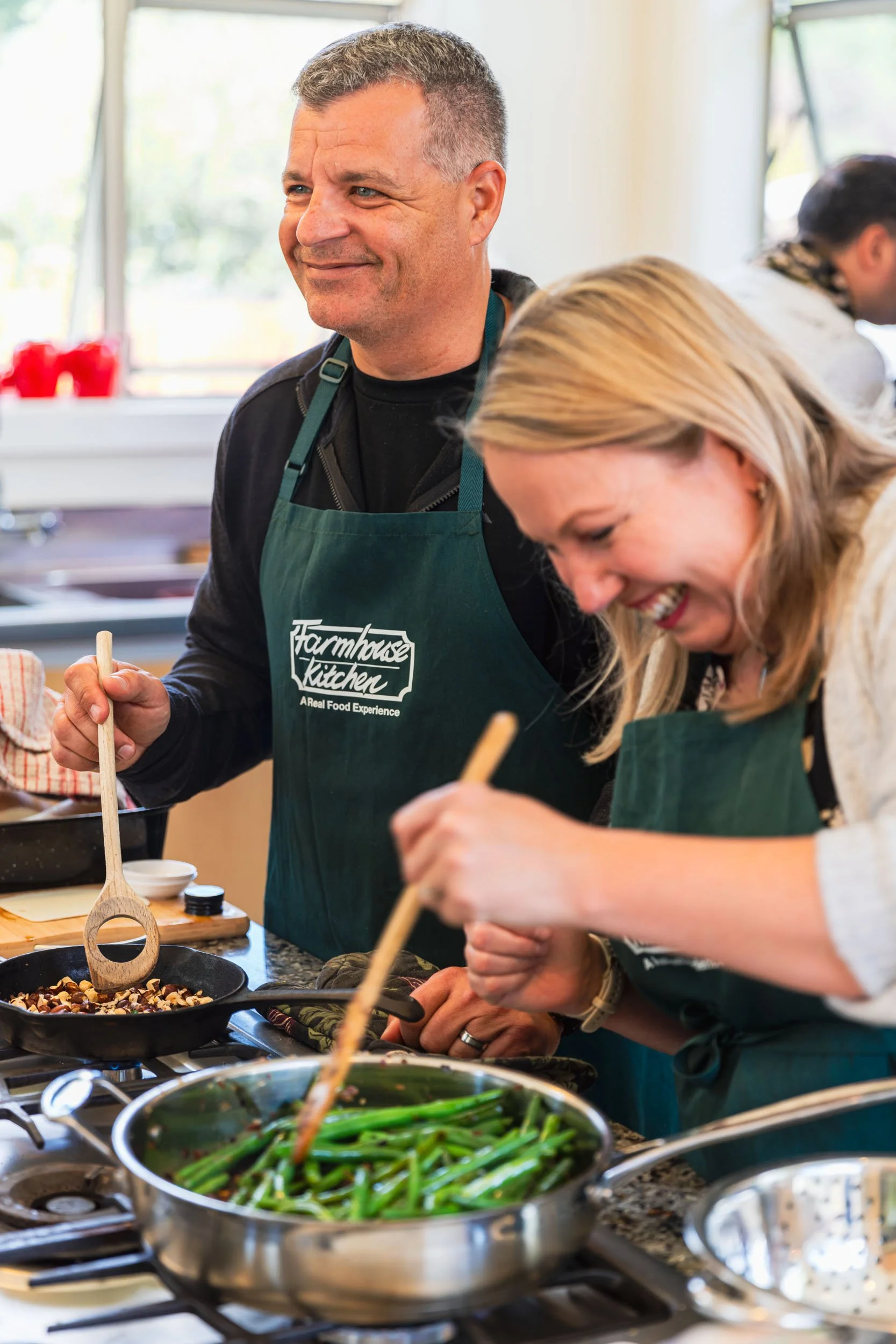 Two people cooking in a kitchen with ovens, wearing aprons, one man smiling and a woman focused on preparing food, with ingredients and cooking utensils on the counter.