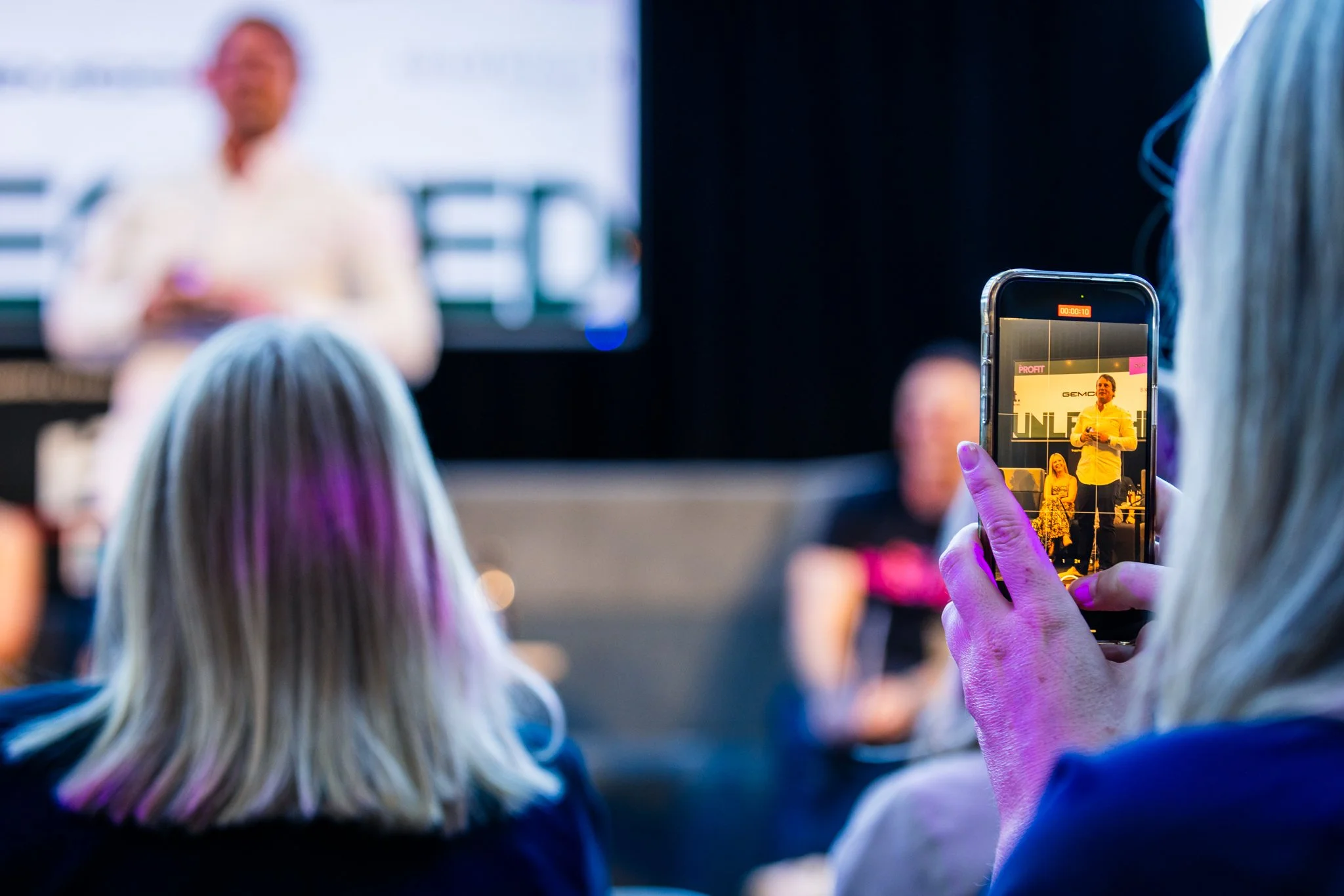 A woman taking a photo of a speaker presenting at an event with her smartphone, focusing on the speaker on the stage.