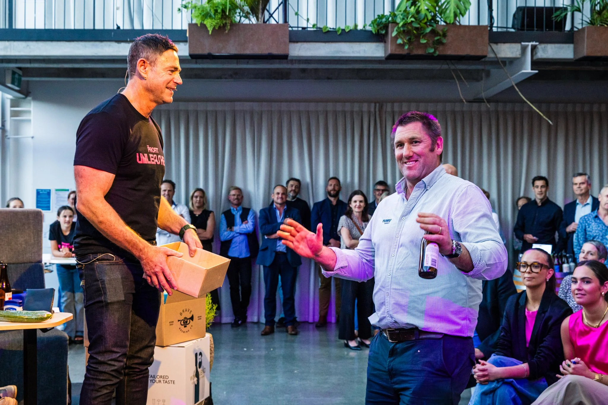 Two men are engaging in a friendly conversation at an indoor event with a group of people watching. One man, on the left, is wearing a black T-shirt and holding a box. The other man, on the right, is wearing a striped shirt and holding a beer bottle.