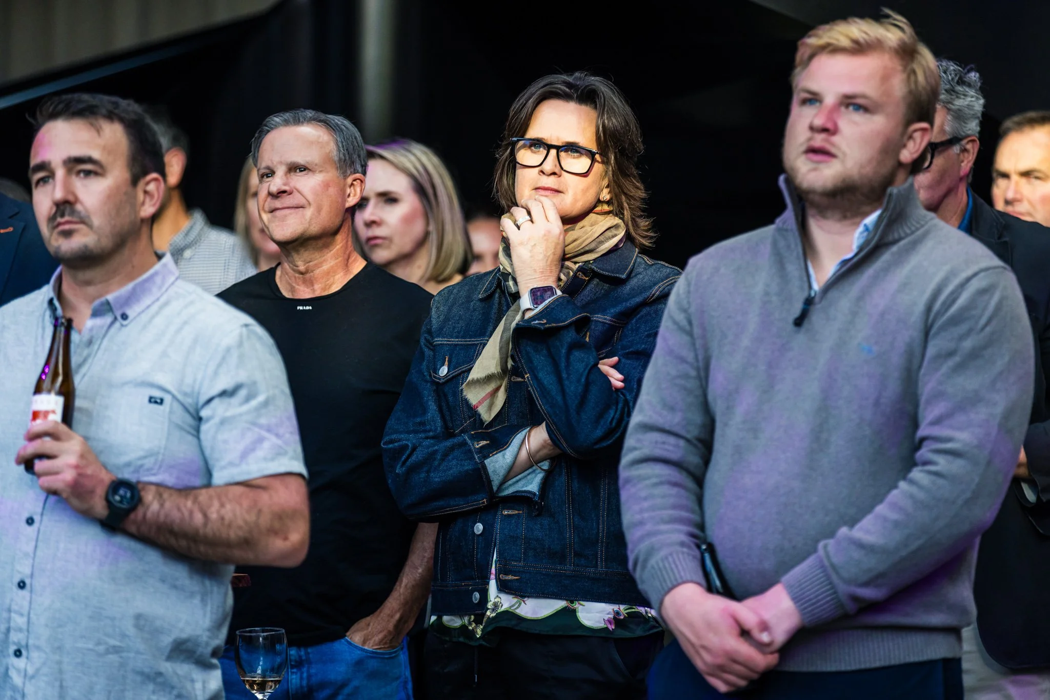 Group of people attending an event, standing and listening intently, with some holding drinks.