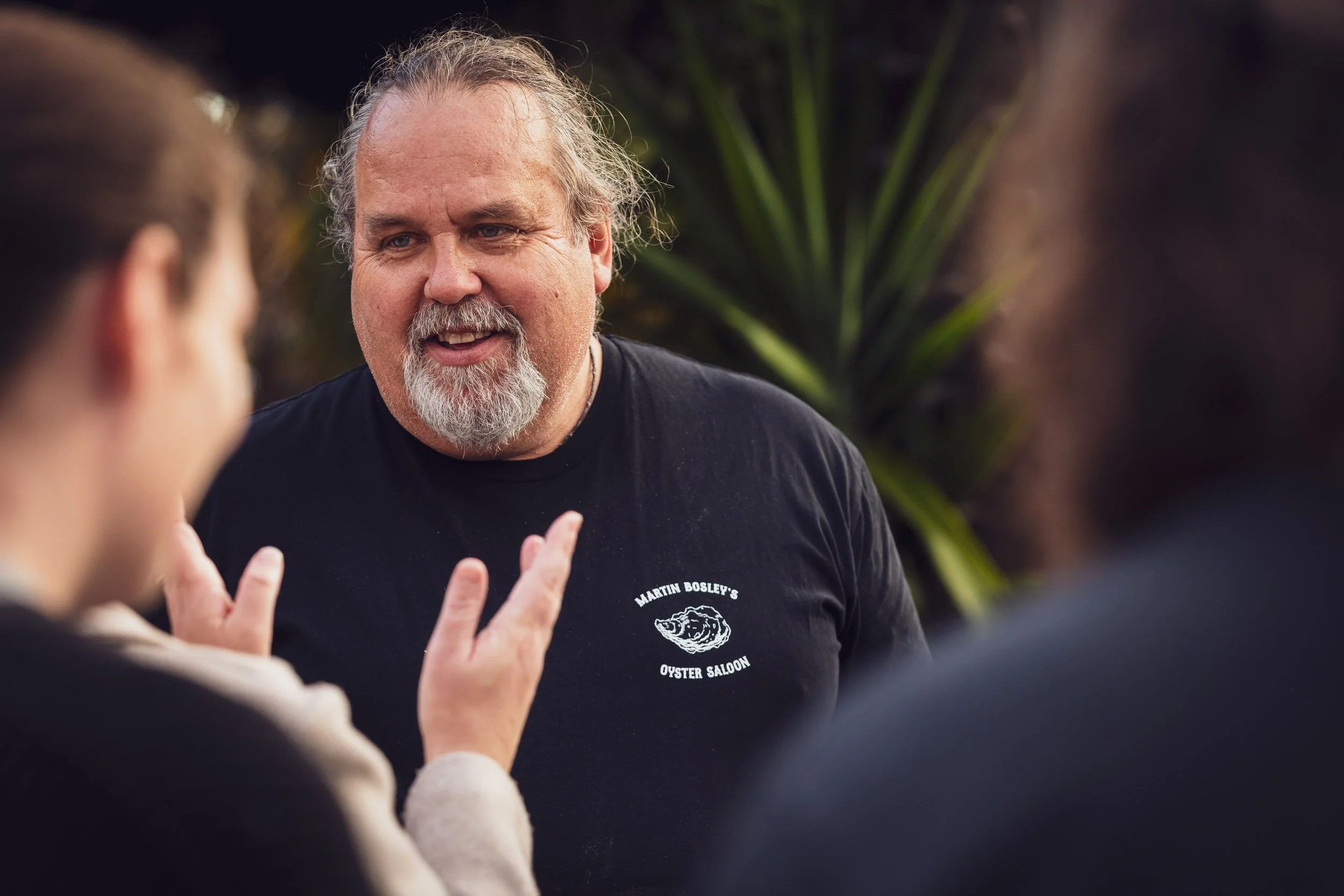 A man with gray hair and a beard, wearing a black T-shirt with white text and an oyster logo, talking and gesturing with a group of people outdoors.