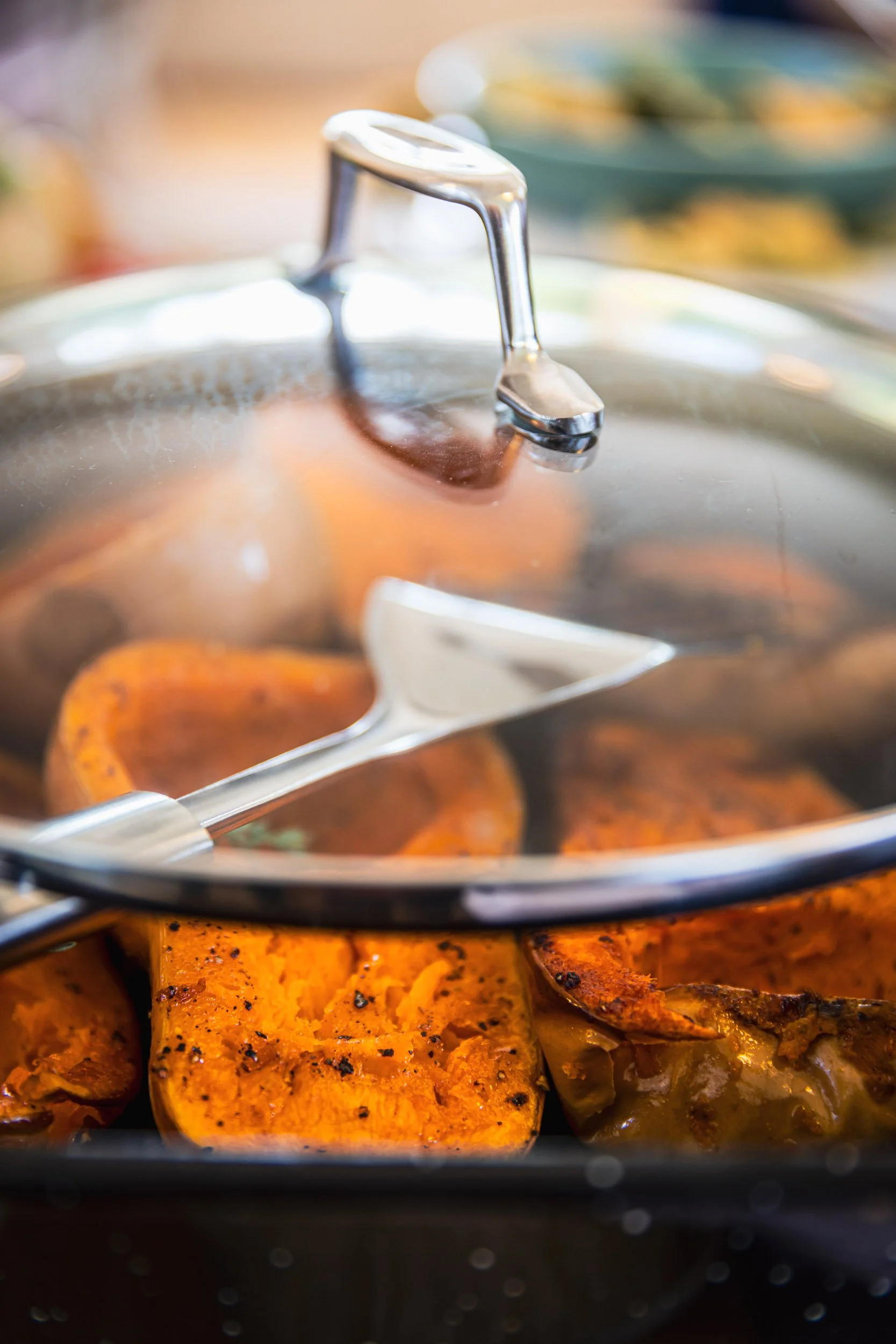 Close-up of a stainless steel lid on a dish containing roasted sweet potatoes, with a slotted spatula resting on top.