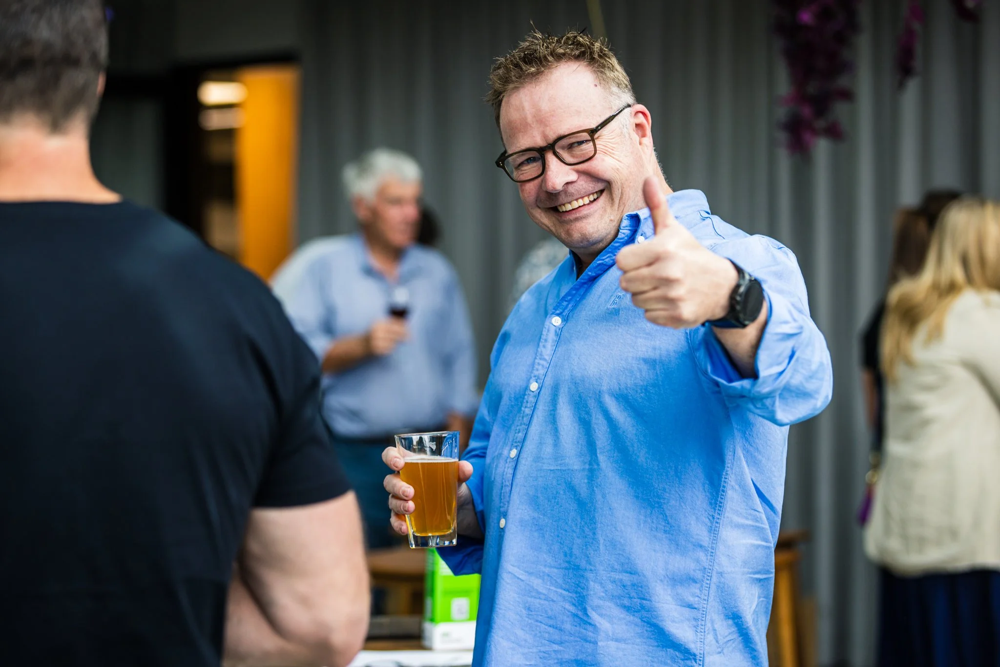 A man in a blue shirt and glasses giving a thumbs-up and holding a glass of beer at a social gathering.
