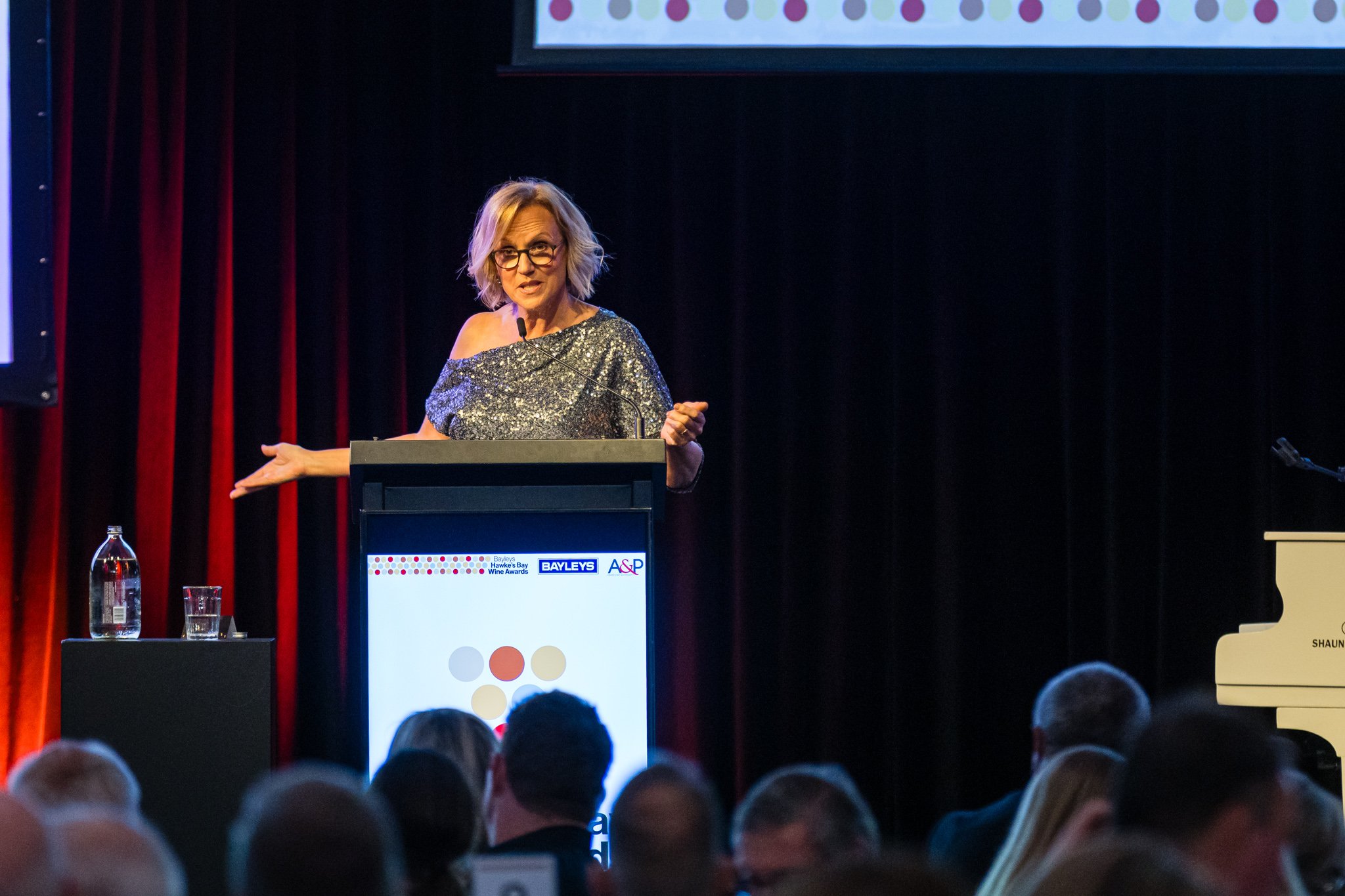 Woman with blonde hair and glasses giving a speech at a podium on stage during an awards event, with a microphone, a water bottle, and a glass on a small table to her left, and a white piano on her right, audience members in the foreground, and a scr