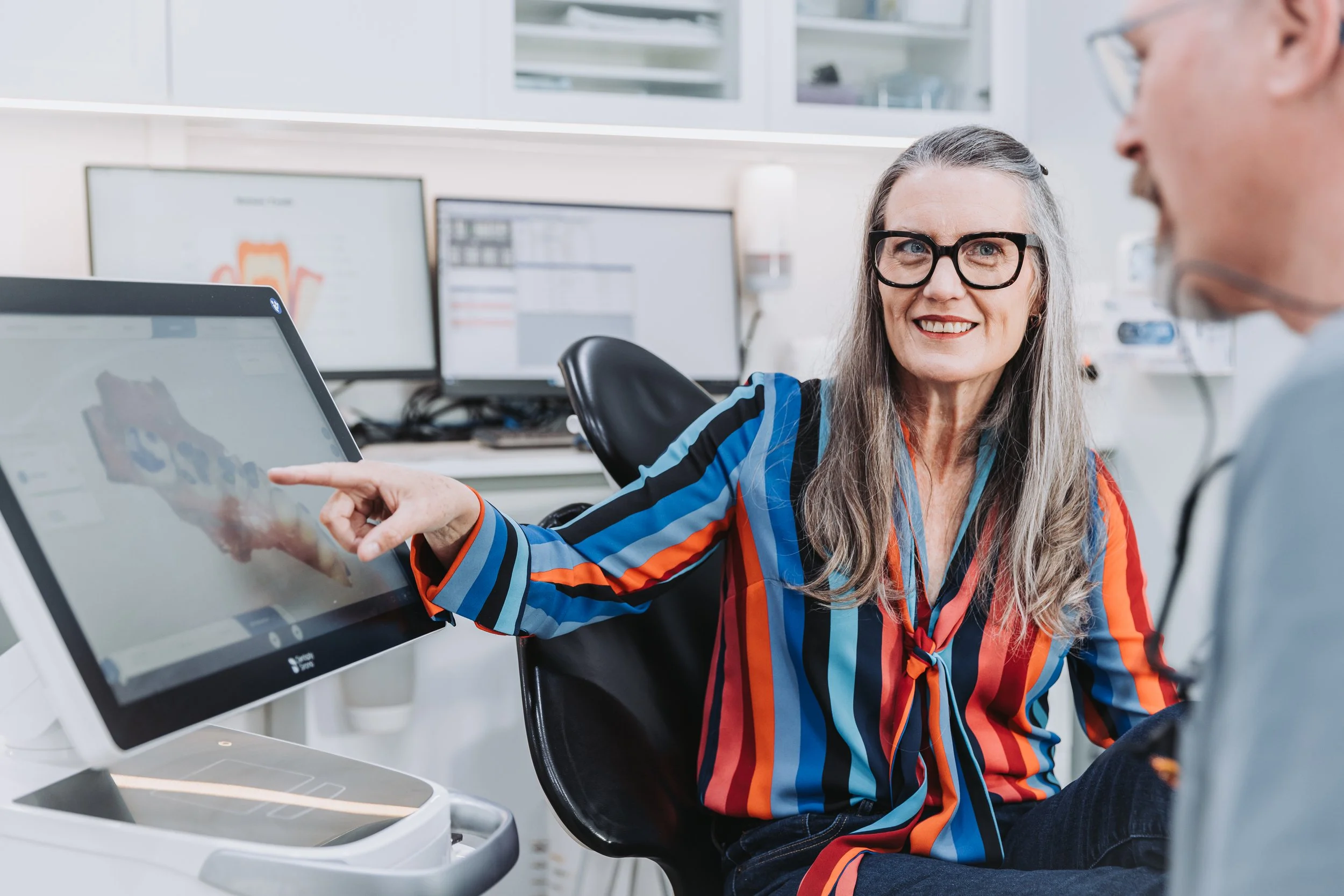 A woman with glasses and long gray hair smiling and pointing at a touchscreen monitor in a medical or dental office, with a man sitting nearby.