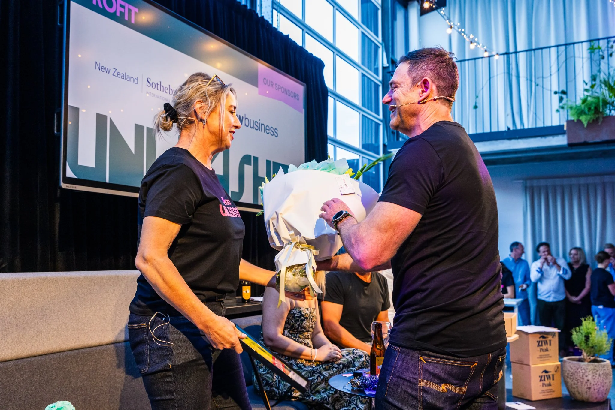 A man is giving a woman a bouquet of flowers on a stage during an event. The woman appears to be receiving recognition or an award. There are several people in the background, with some seated and several others standing, in a well-lit indoor venue w