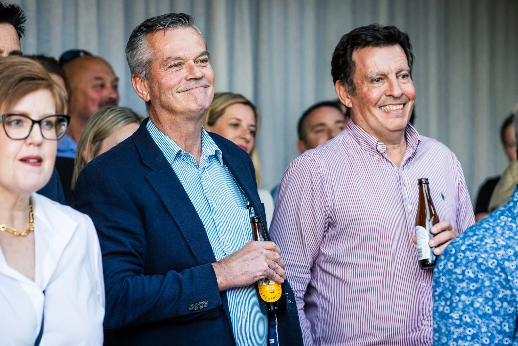 Group of people attending a social event, some holding beer bottles, smiling and engaging in conversation, with a focus on two men in the center.