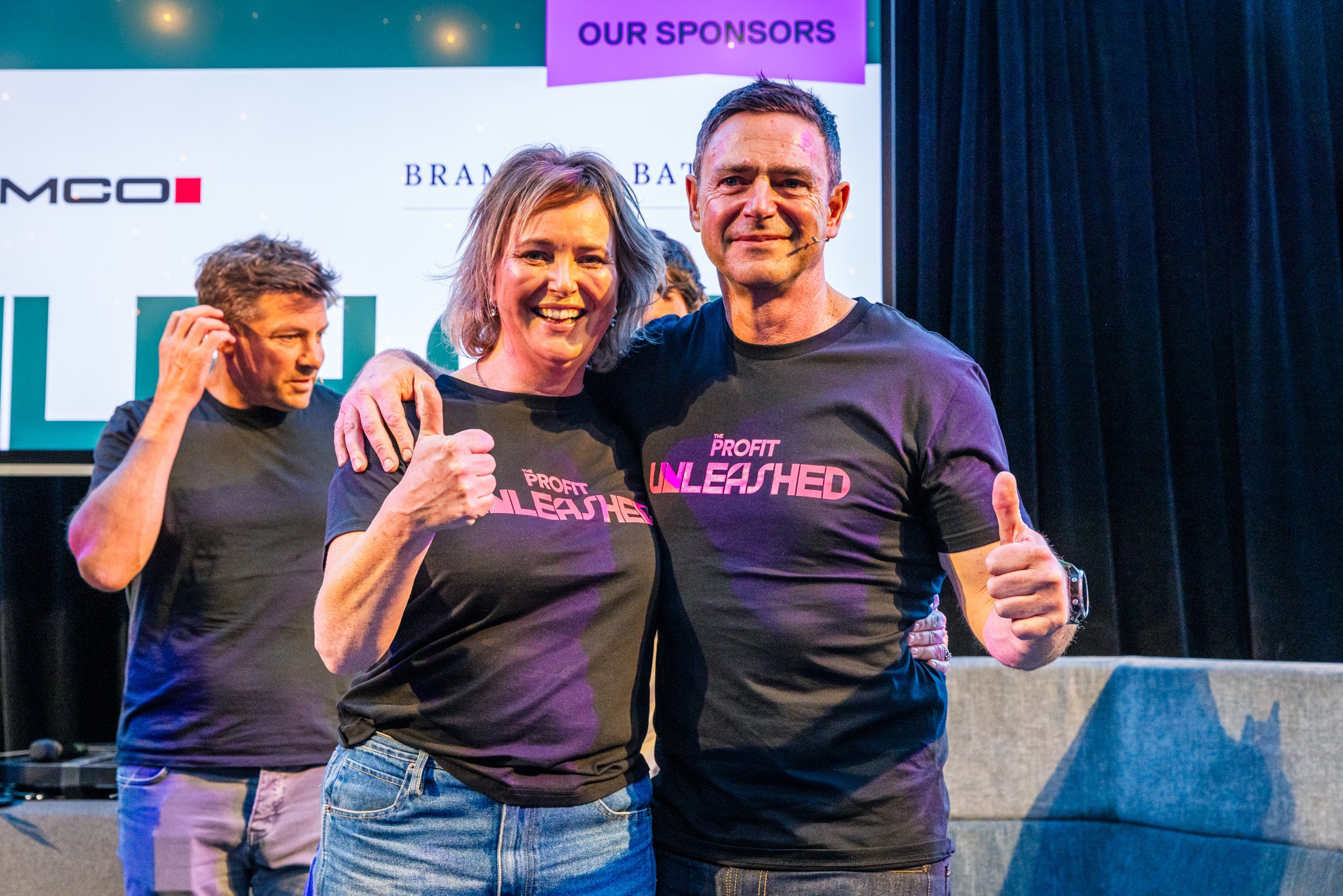 Three people on stage at an event, two in the foreground smiling and giving a thumbs up, wearing matching black shirts with pink text that reads "Profit Unleashed," and a man in the background adjusting his hair with a serious expression.