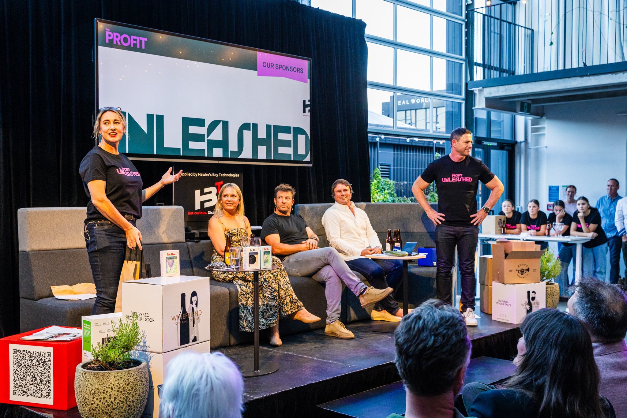Panel discussion at a conference with five speakers, three sitting and two standing, in front of a large screen displaying "UNLEASHED," with audience members visible in the foreground and background.