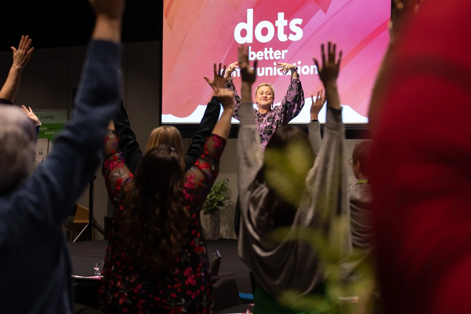 A woman leading a seated audience in a group activity or exercise in a conference room, with a large screen behind her displaying the words "dots better communication." The audience members have their hands raised and are participating actively.