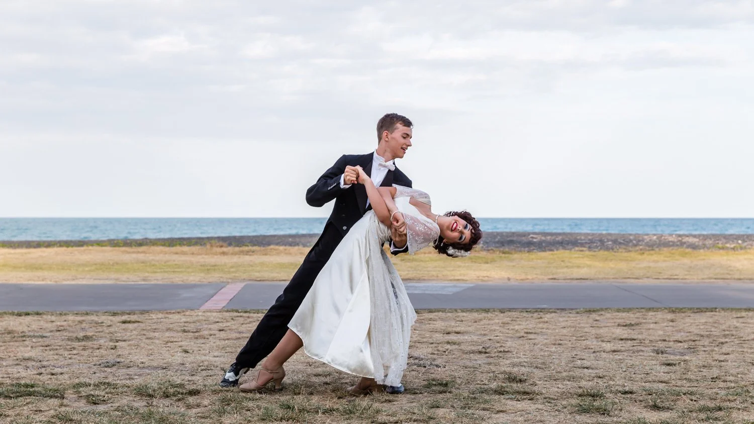 A couple dancing outdoors near the ocean, with the man dipping the woman in a white dress, both smiling.