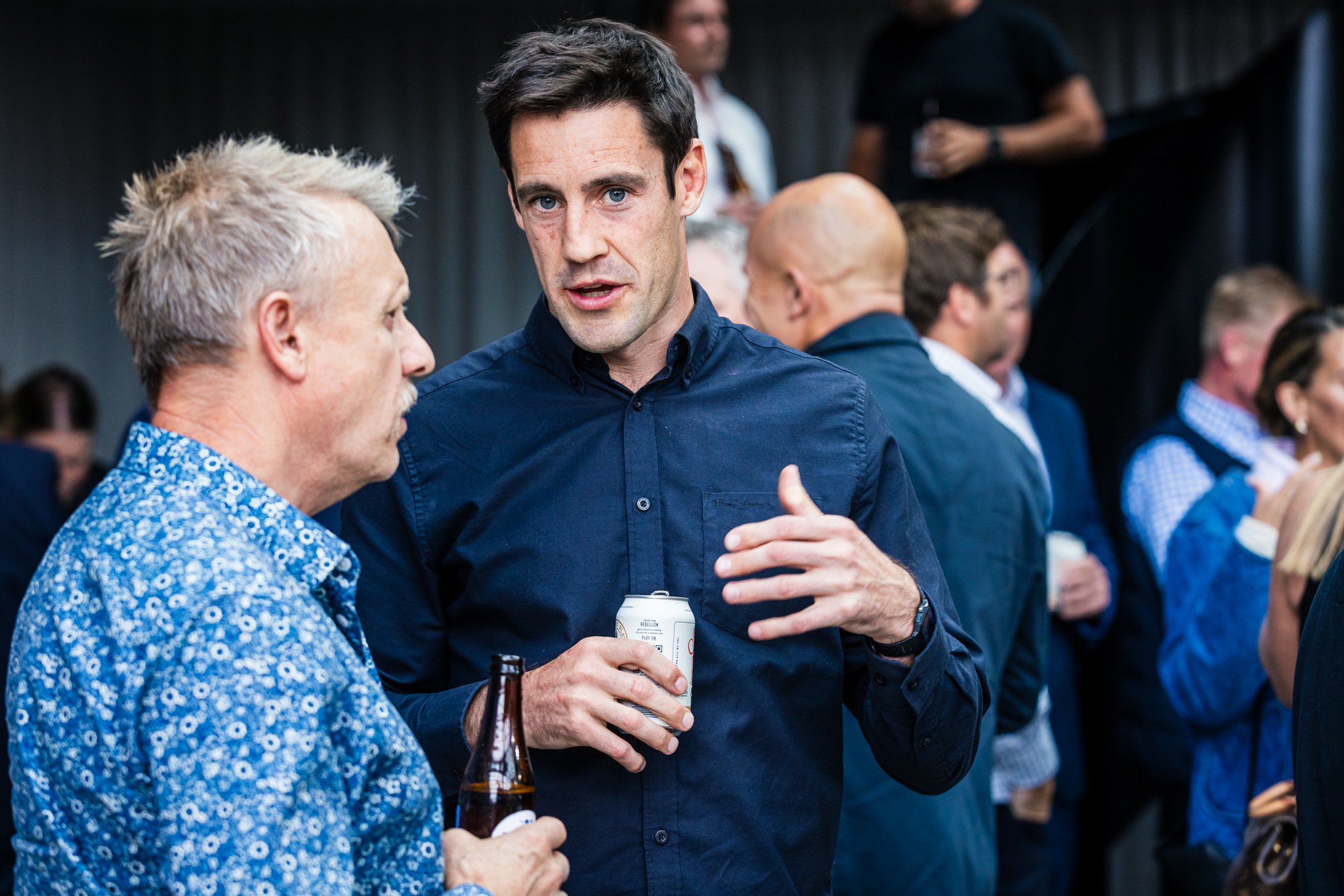 Two men engaged in conversation at a social event, one holding a beer bottle and the other holding a can, with a group of people in the background.