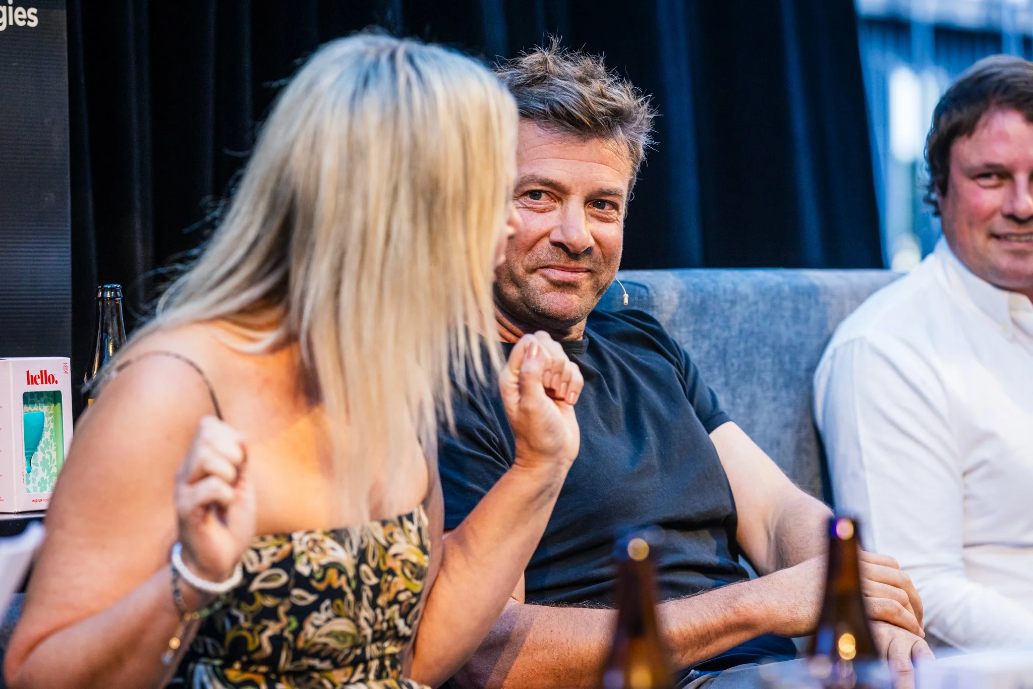 Three people sit on a sofa at a social gathering, engaging in conversation, with bottles and a Hello! gift box on the table.
