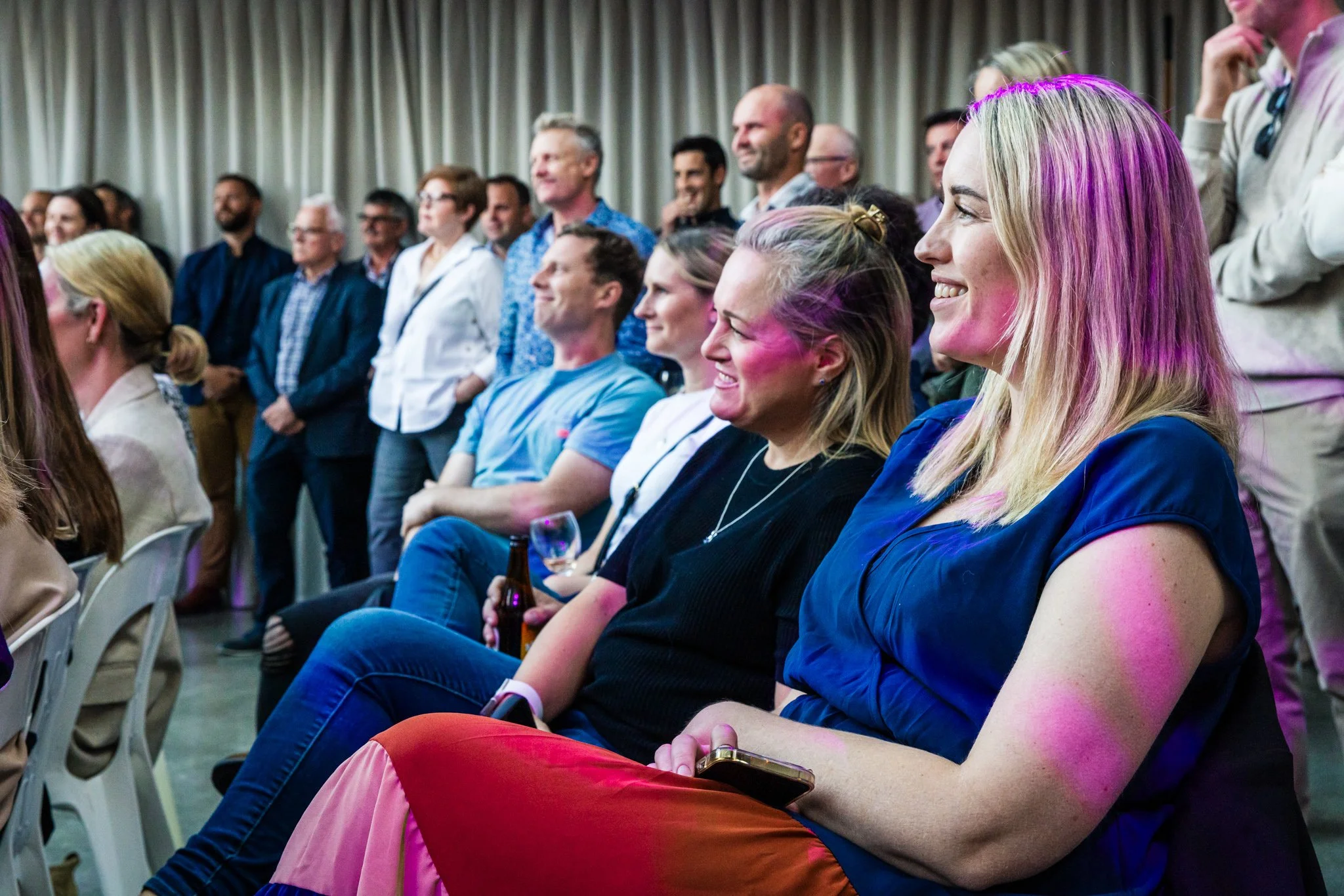 People sitting and standing in a conference room, smiling and watching a presentation or event.