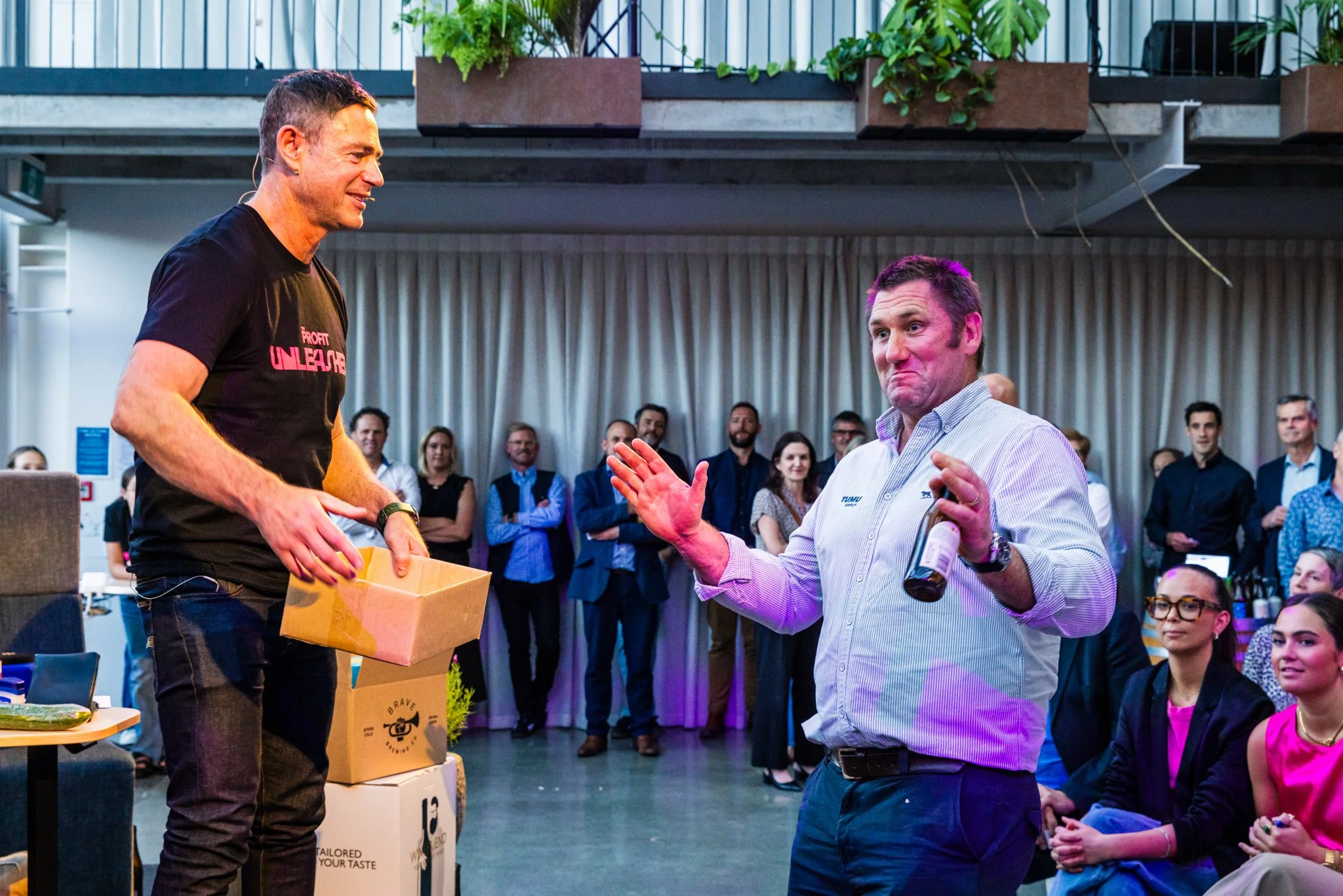 A man in a black t-shirt standing on a stage with a smile, holding a box, receiving a beer from another man in a striped shirt holding a beer bottle while making a gesture with his hand at a gathering with an audience of diverse men and women in busi