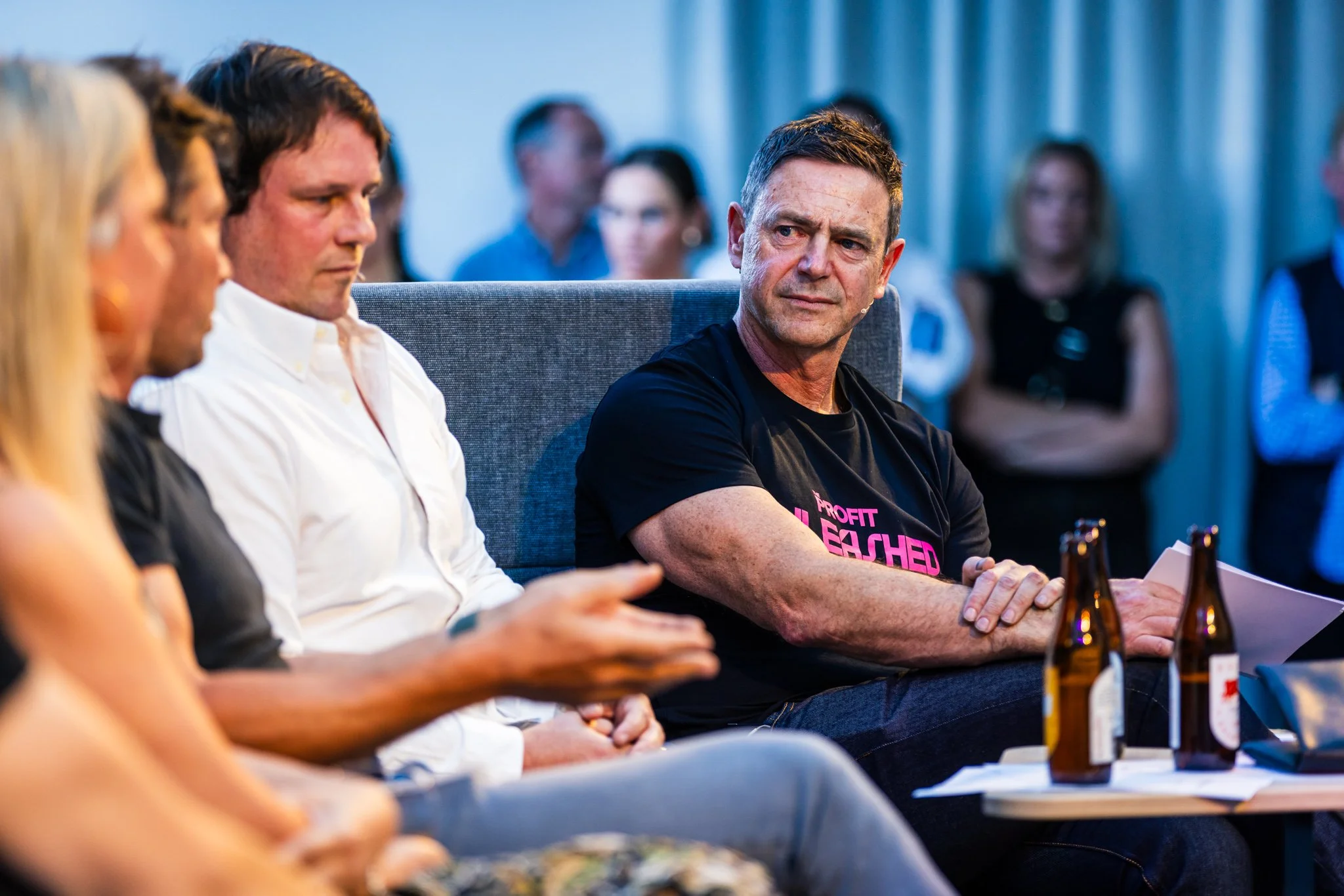 A group of people sitting and listening in a meeting room, with one man in a black T-shirt prominently in focus, wearing a T-shirt that says 'Profit Unleashed.'