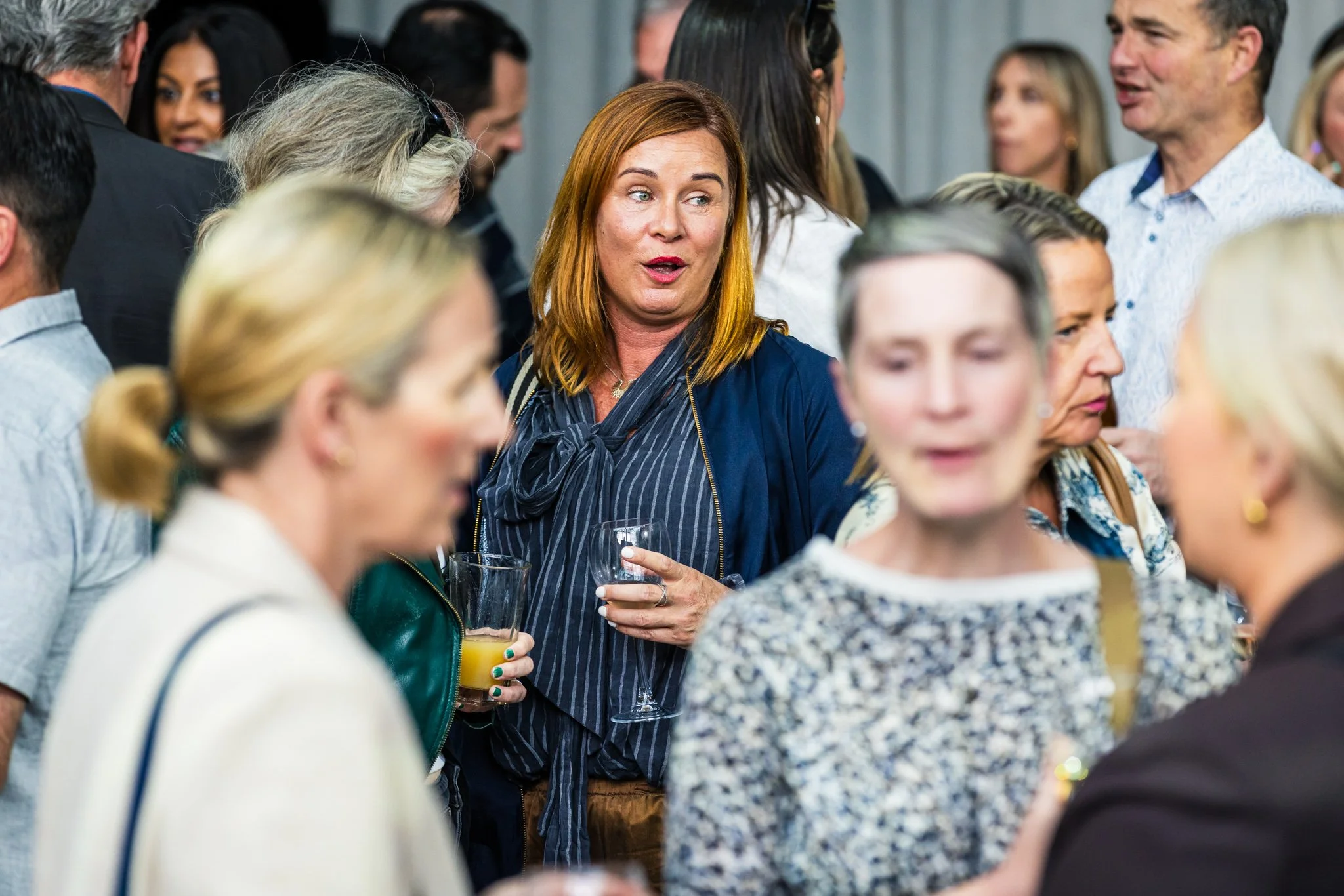A group of people at a social gathering, talking and holding drinks, with a woman with red hair at the center.