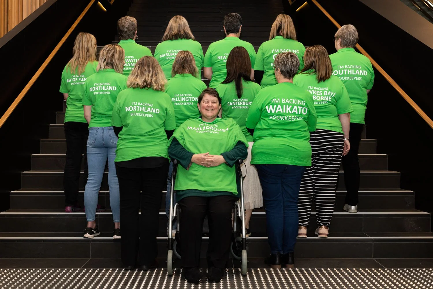 Group of people wearing bright green T-shirts with various messages supporting different regions, standing on a staircase indoors, with a person in a wheelchair smiling at the camera in the front center.