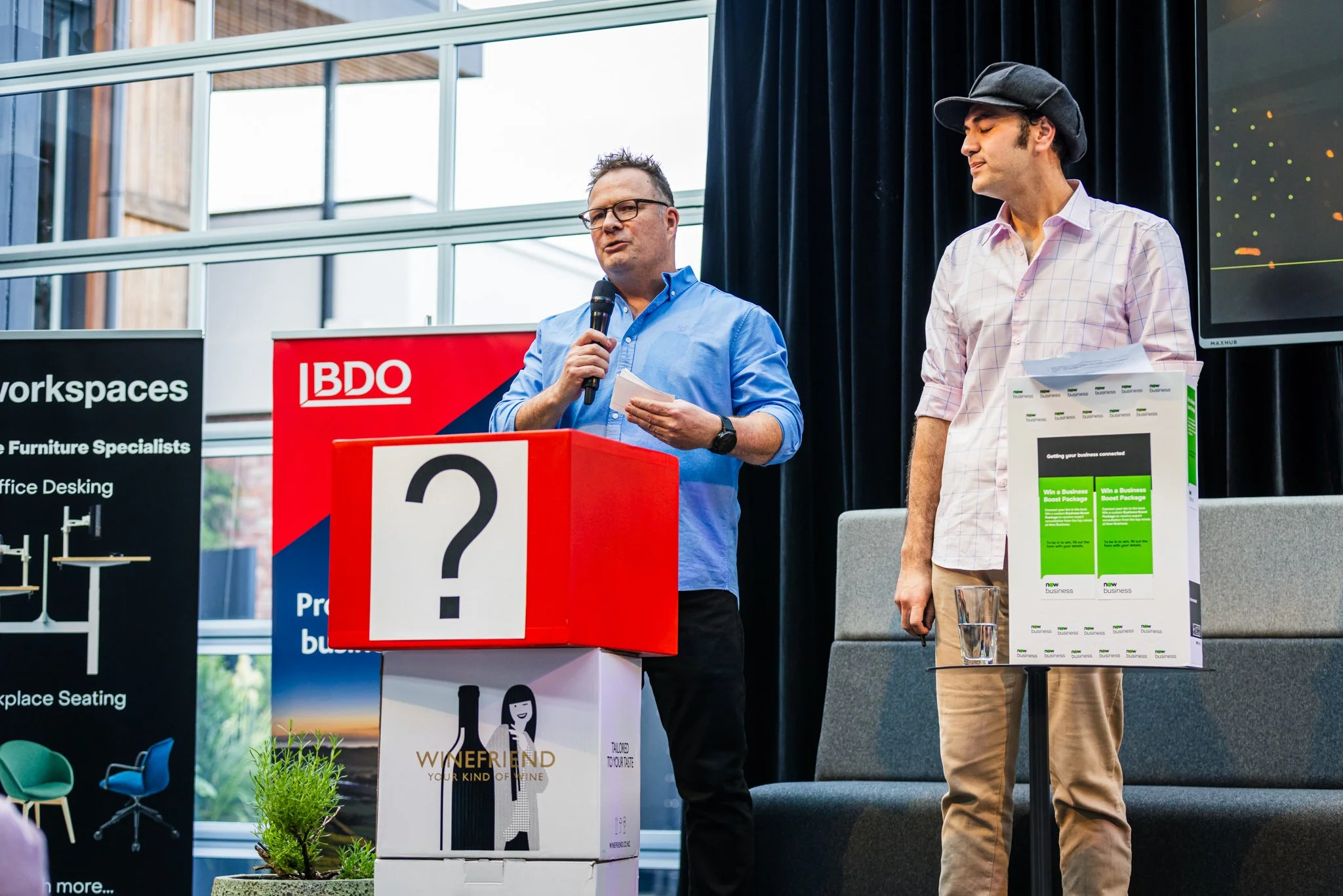 Two men on a stage participating in a presentation or event, with one holding a microphone and reading from notes, the other standing nearby. There are promotional banners, boxes, and a large screen in the background.