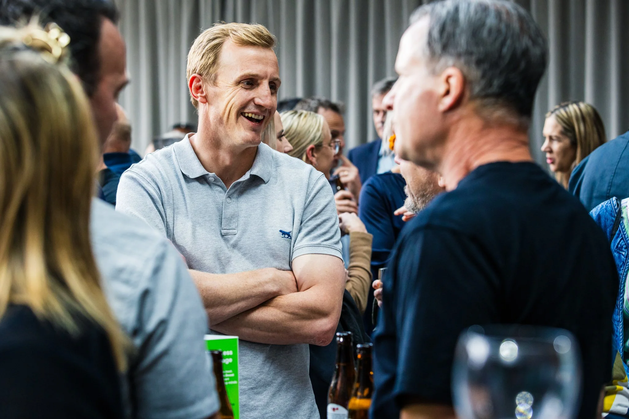 A group of people socializing at a gathering with a man in a light gray polo shirt smiling and crossing his arms, engaged in conversation with a man in a black shirt. In the background, others are talking and holding drinks.