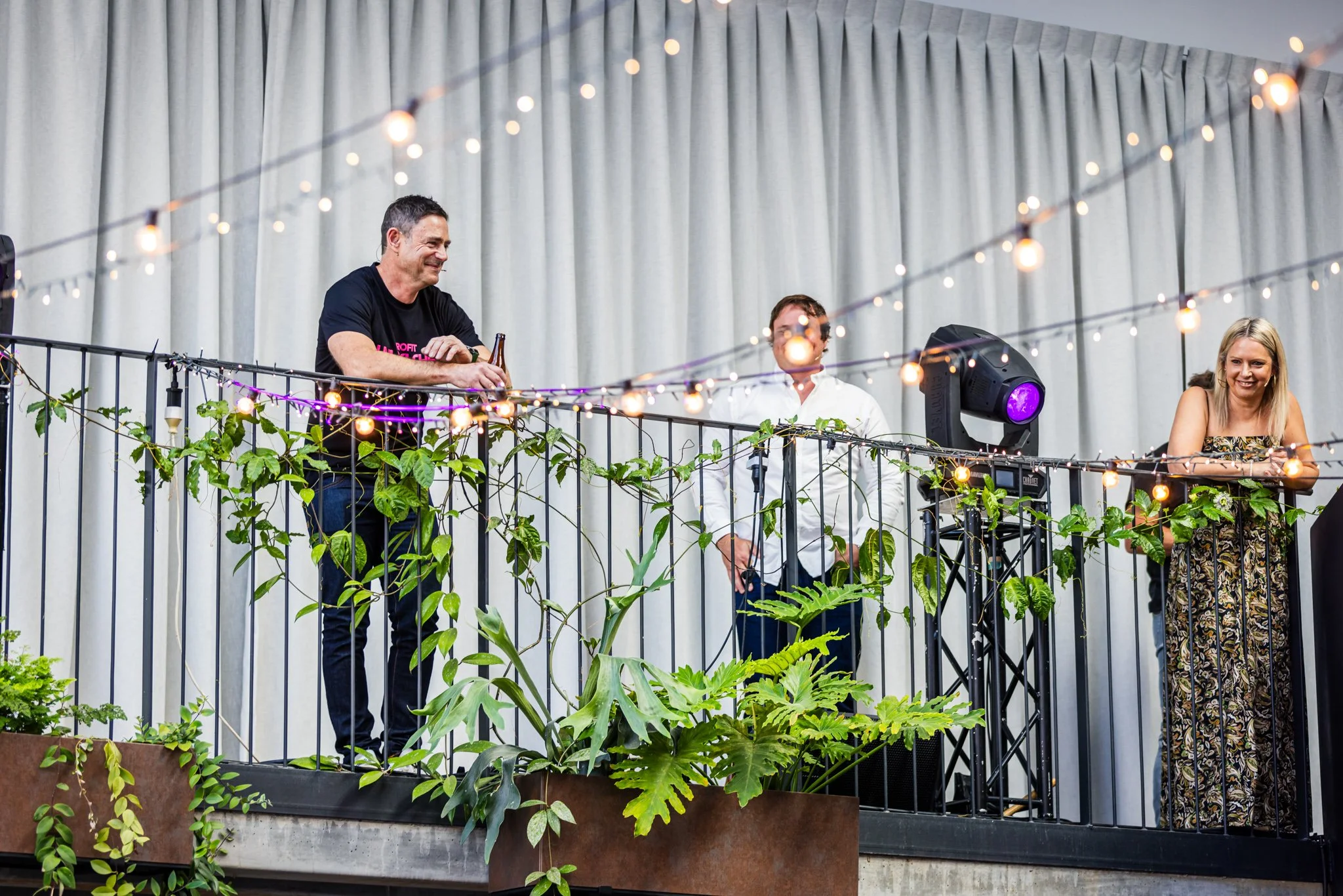 Three people standing on a decorated balcony with string lights and greenery, engaged in conversation or listening to music, against a backdrop of a white curtain.