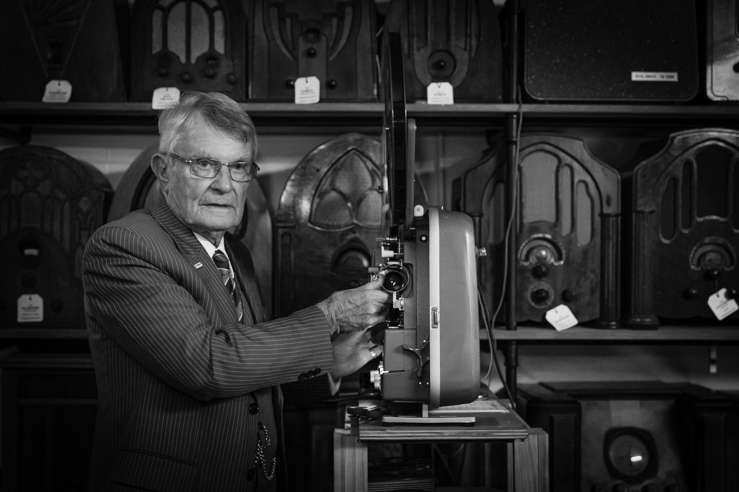 An elderly man in a pinstripe suit and glasses operating a vintage film projector in a room filled with older radios.