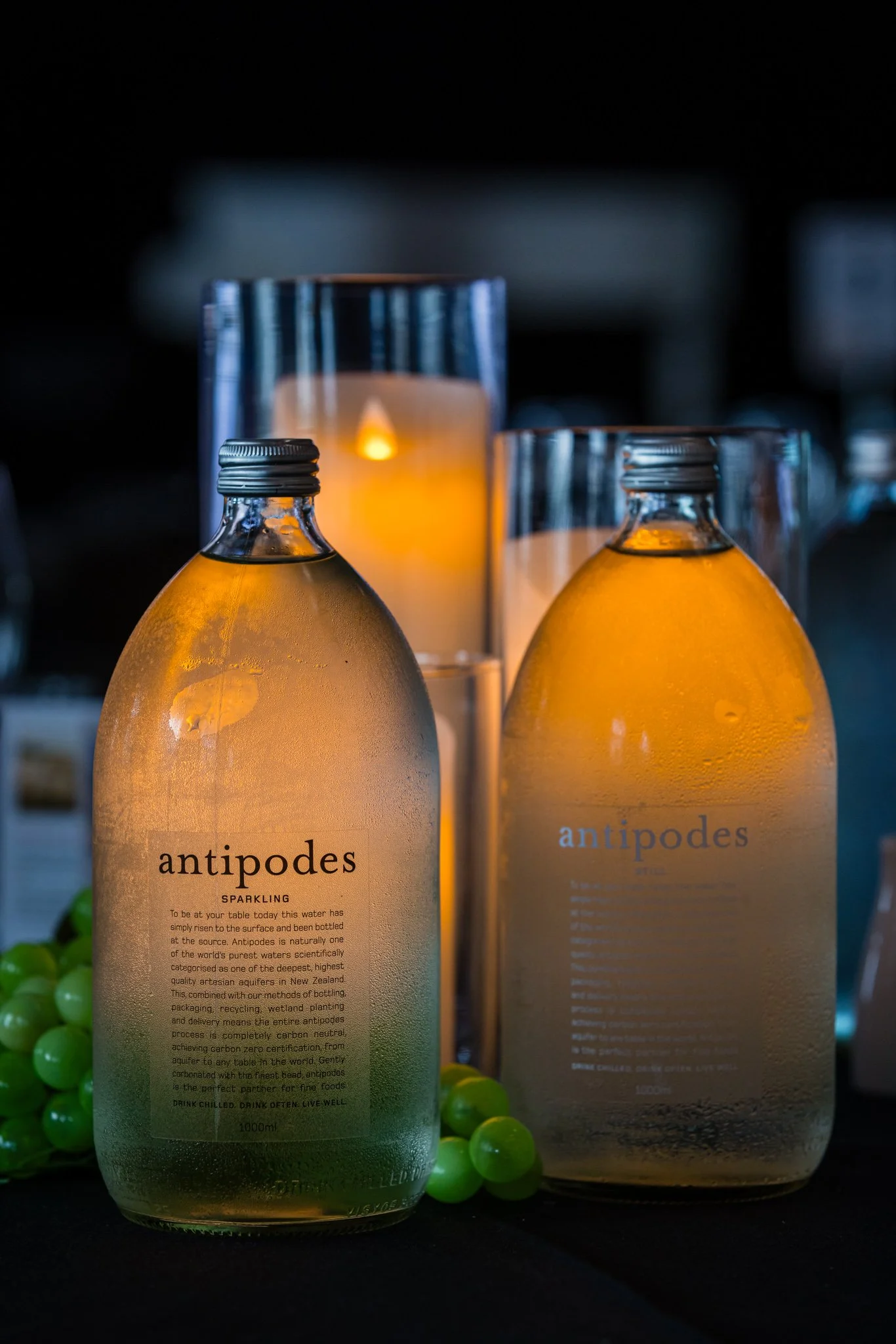 Two bottles of Antipodes sparkling water in front of lit candles, with green grapes beside.