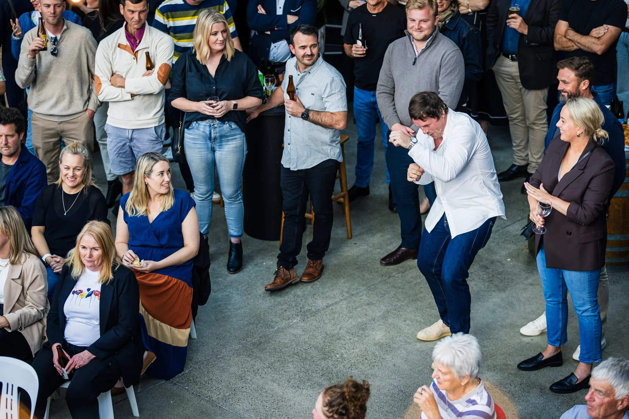 Group of people at a social gathering, some standing, some seated, with one woman dancing and others holding drinks and chatting.