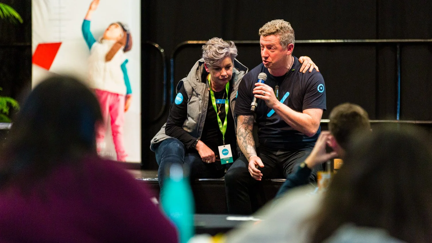 A man with tattoos holding a microphone and talking to an audience, with a woman sitting next to him leaning in, in a conference setting. Blurred audience members are in the foreground.
