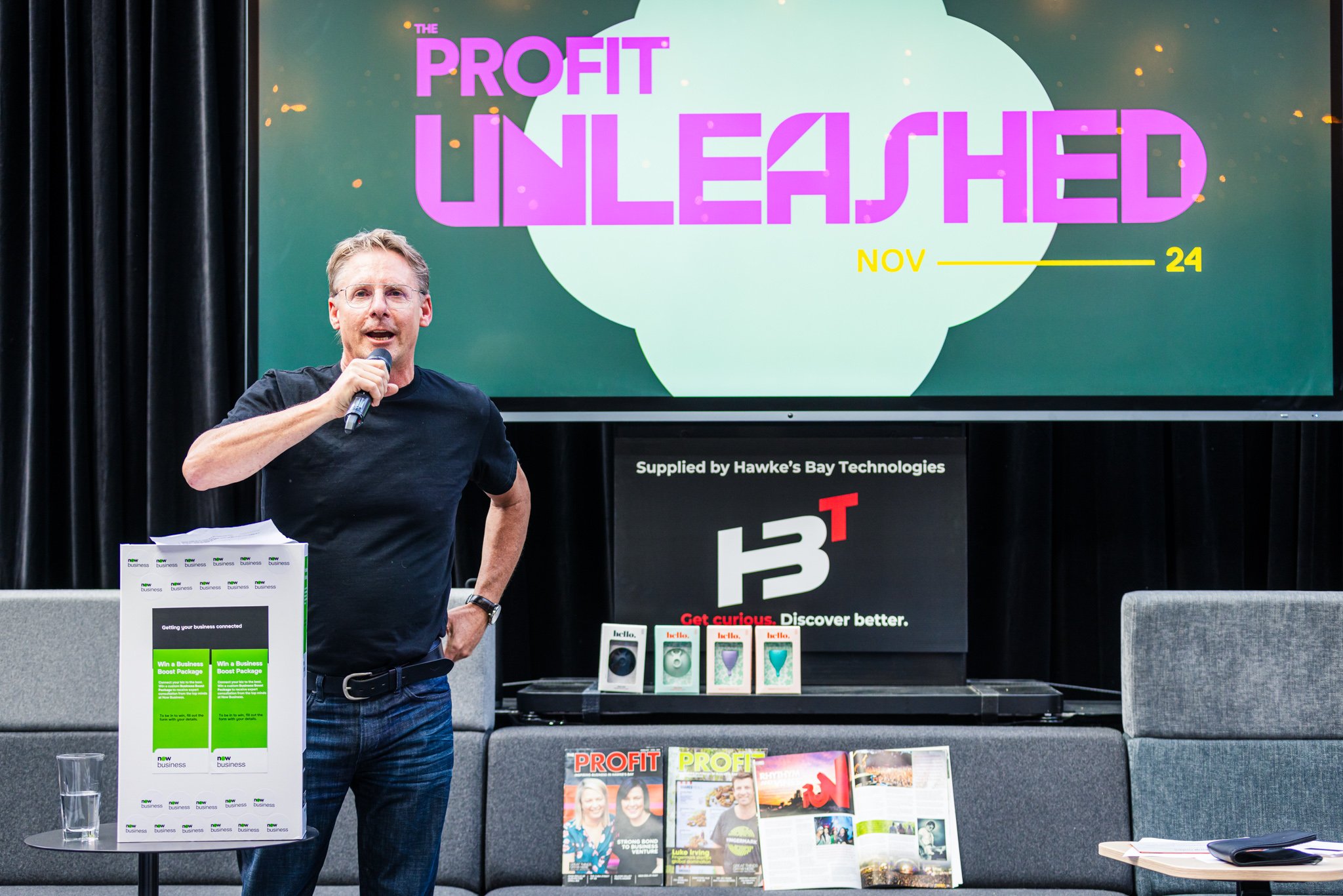A man holding a microphone and wearing a black t-shirt, speaking at an event with a large screen behind him displaying the text 'The Profit Unleashed, Nov 24'. There are promotional materials and magazines on a table and shelf, with a black curtain b