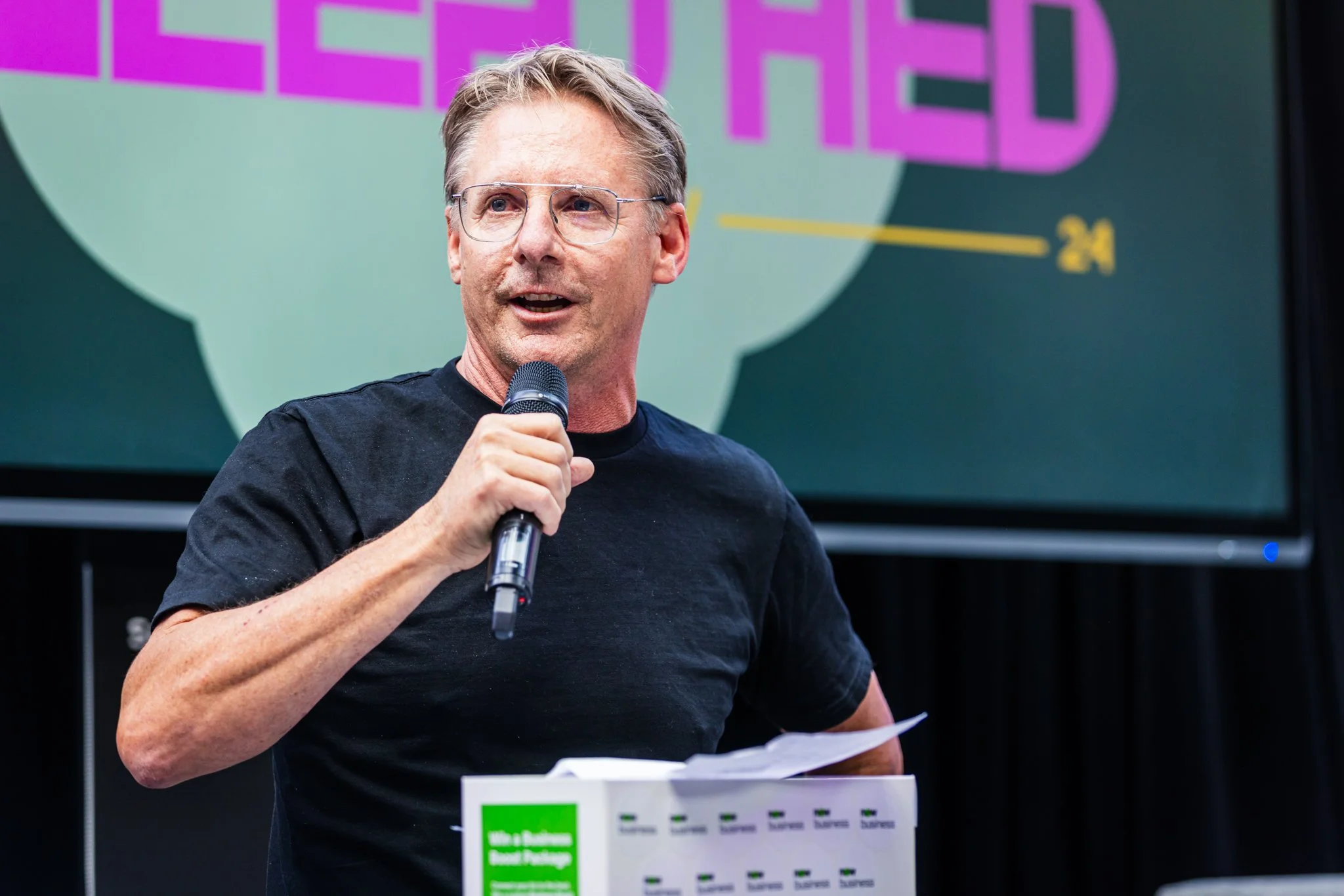 A middle-aged man with glasses and short light brown hair, holding a black microphone, speaking at an event with a colorful background that partially reads 'GREEN FED' and has some yellow text, while standing behind a podium with a green label.