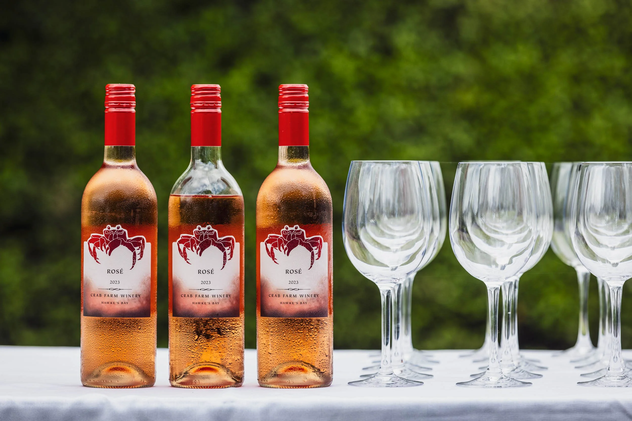 Three bottles of rosé wine labeled from Crab Farm Winery with empty wine glasses on a white table, outdoors with a green blurred background.
