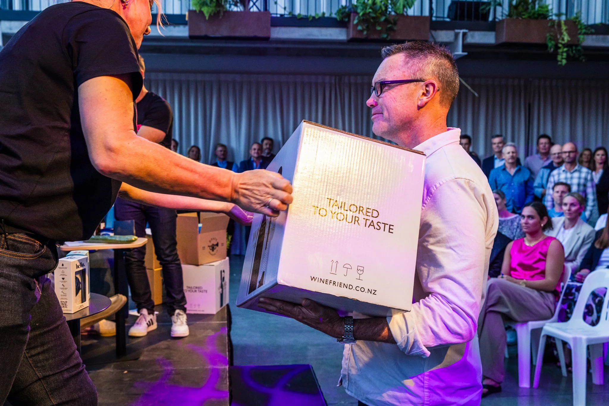 A man in glasses and a white shirt receives a boxed gift from a woman during an event, with an audience seated in the background.