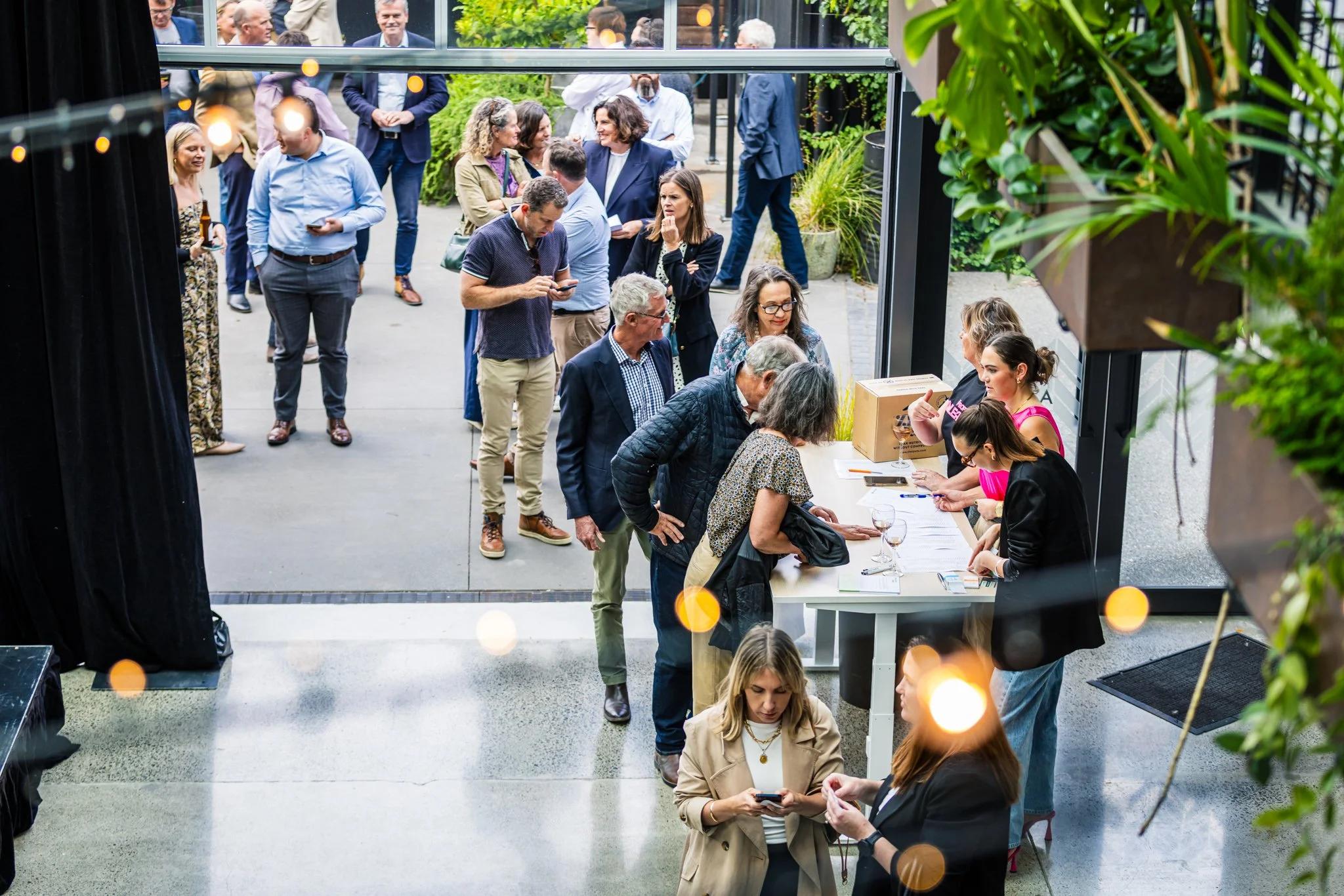 People standing in line at a registration or check-in table inside a modern building with glass doors, with more people mingling outside in a green outdoor area.